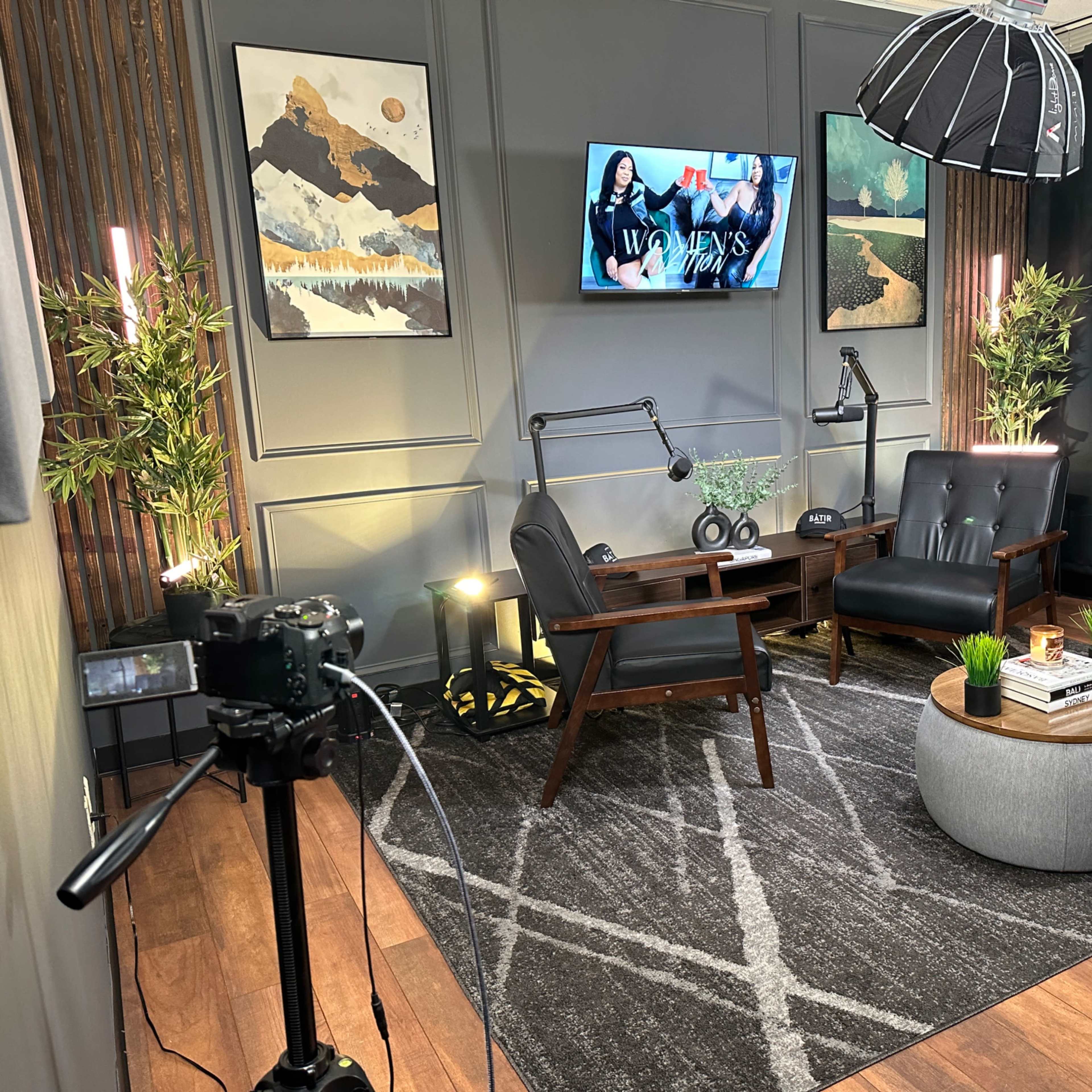 A modern studio setup with two black chairs, a round table, and a large television screen on the wall, surrounded by decor and lighting equipment.