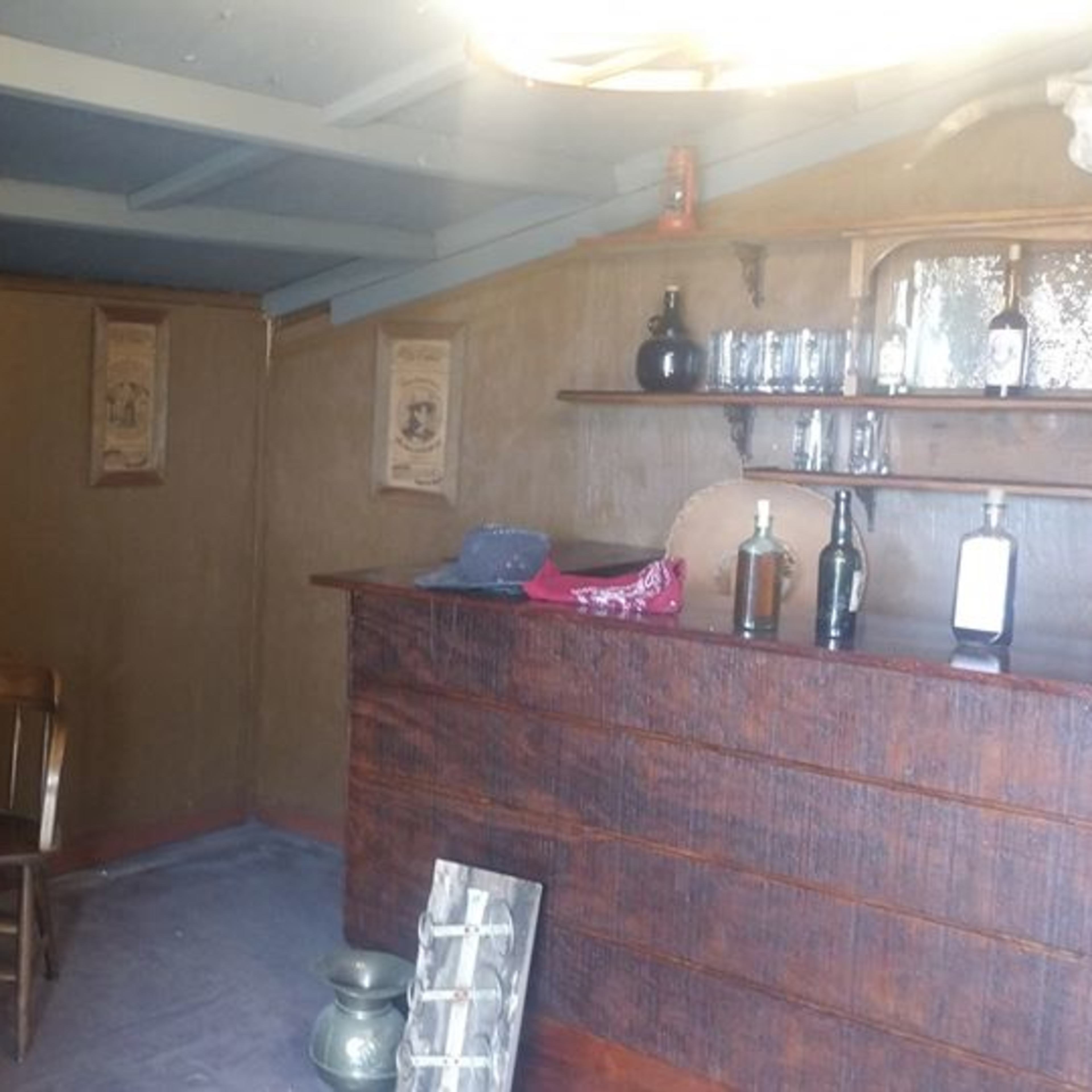 A small, rustic room featuring a wooden table with chairs, a bar with bottles, and period-appropriate decor on the walls.
