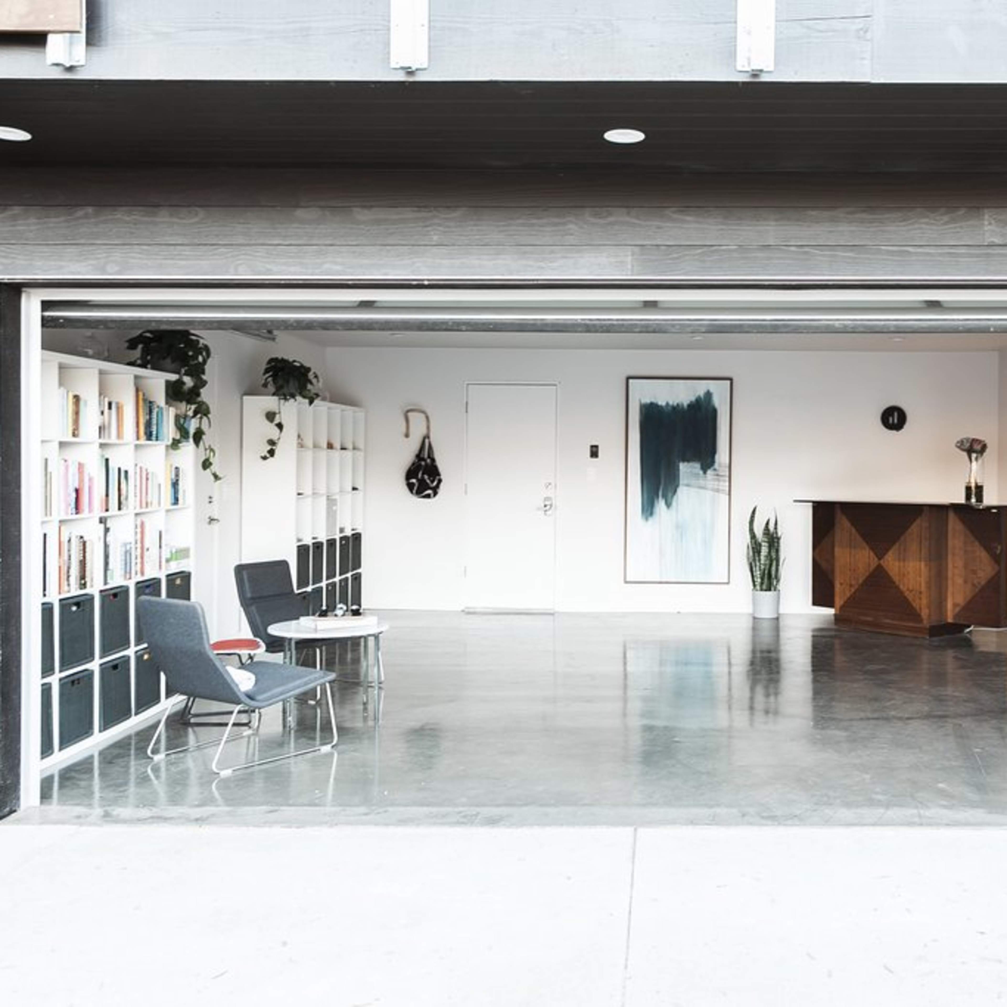 A spacious interior features a garage door open to a minimalist living area with bookshelves, a bar cabinet, and potted plants.