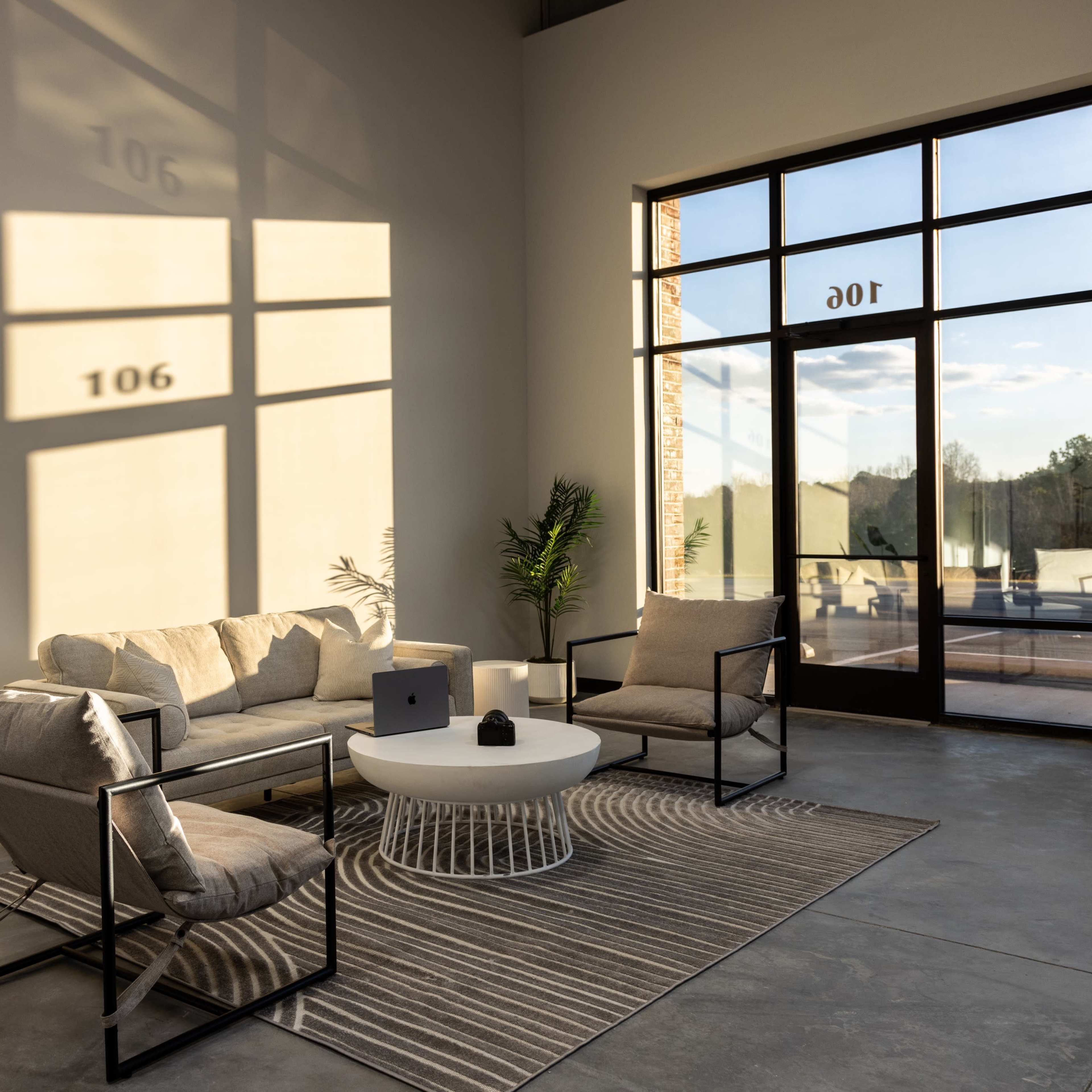 A modern living space features a beige sofa, two chairs, and a round coffee table set on a striped rug, with sunlight casting geometric shadows on the wall.