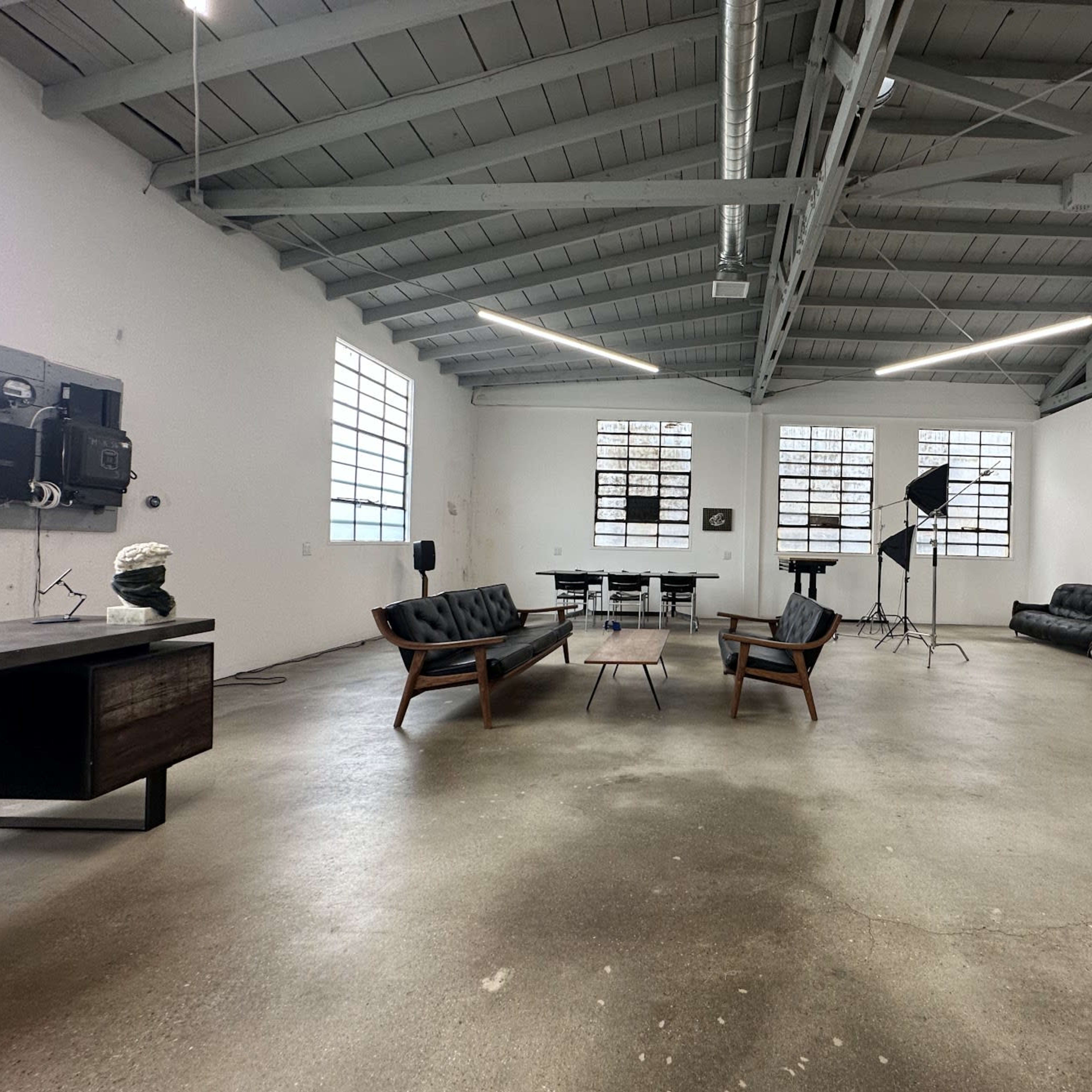 A spacious, industrial-style room with high ceilings, featuring modern furniture including a seating area, dining table, and several large windows.