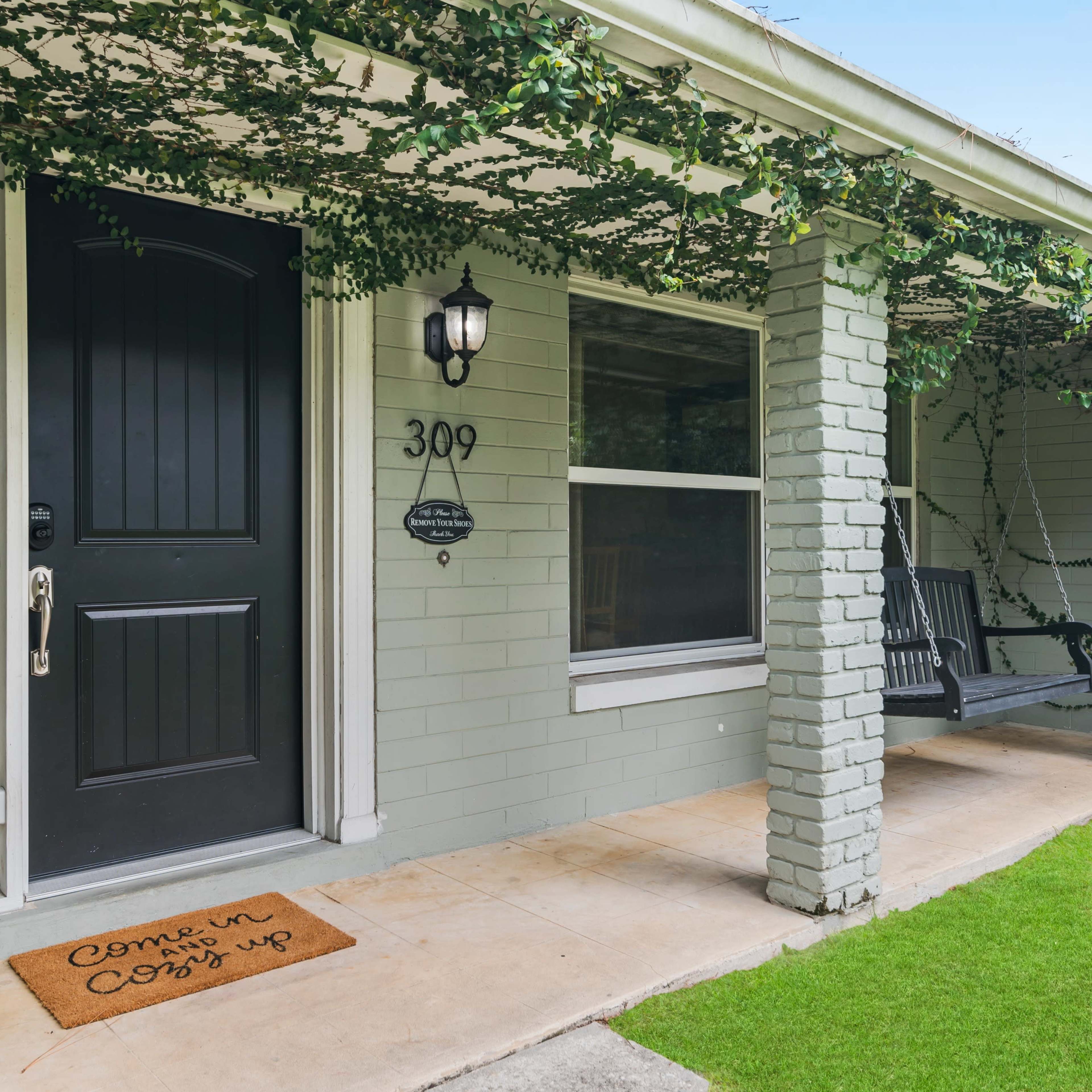 A house entrance with a black front door, a welcome mat, and a porch swing, surrounded by green vines.