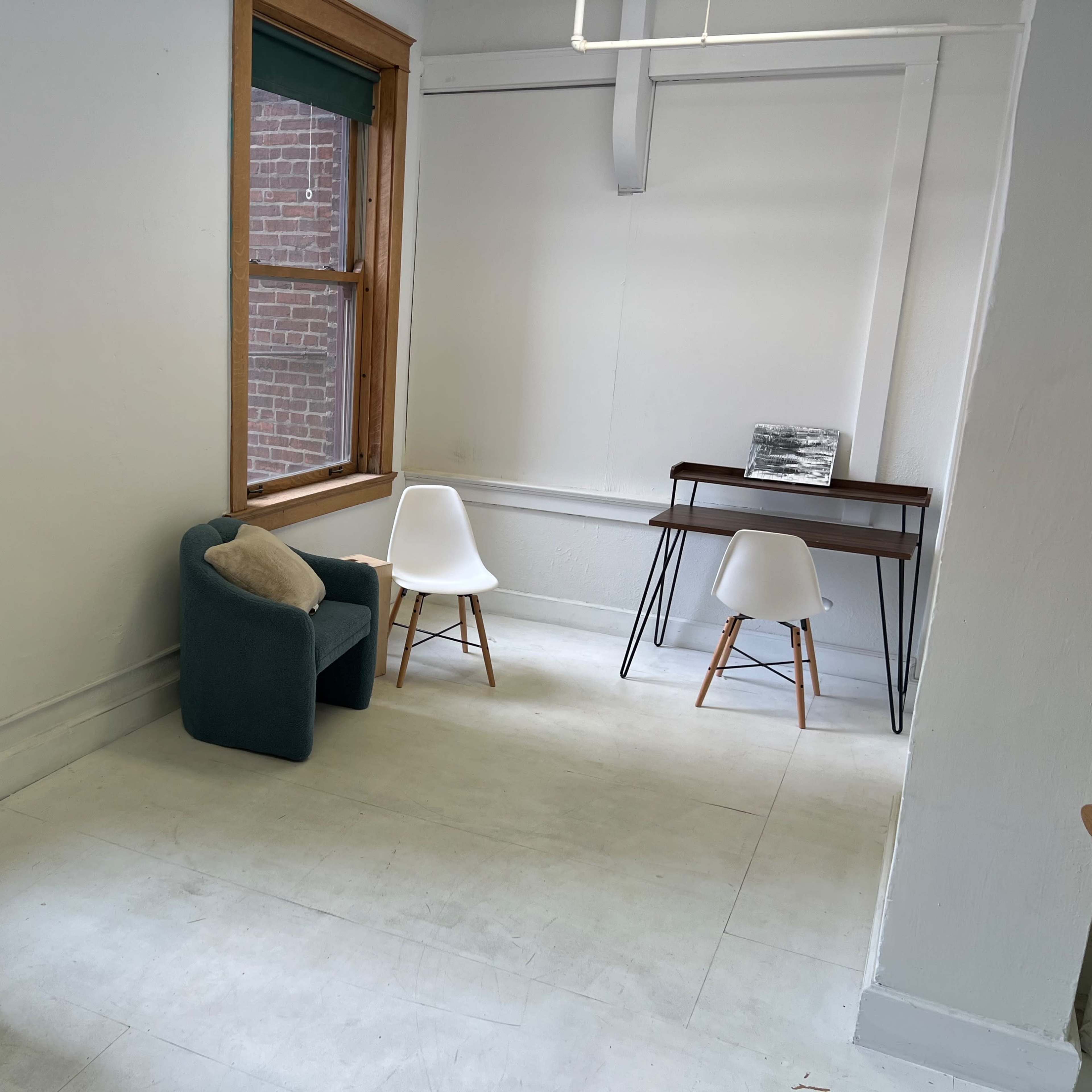 A room with a piano, a teal chair, a small desk, and two white chairs next to large windows.