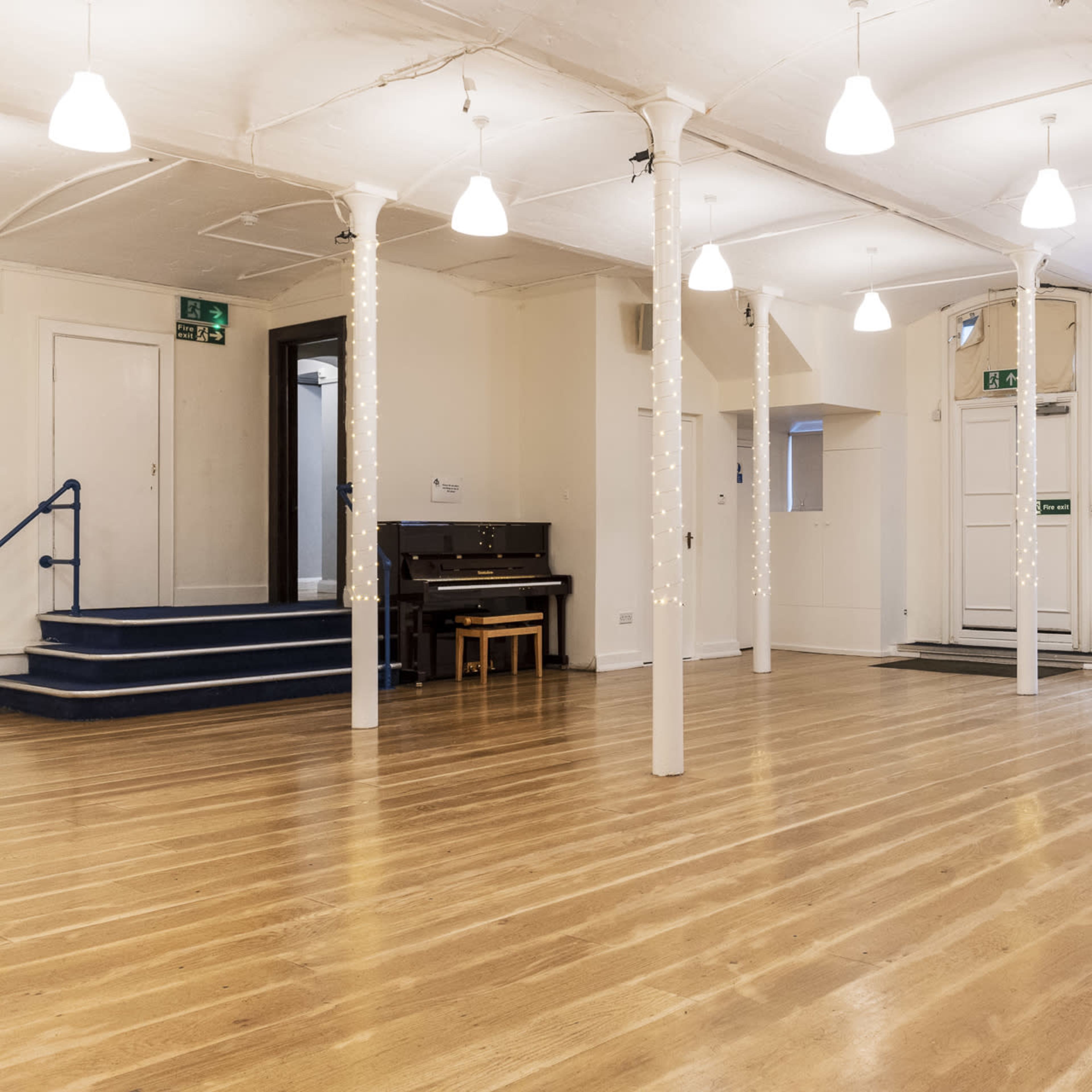A spacious, well-lit room with wooden flooring, white pillars decorated with string lights, and a small piano against one wall.