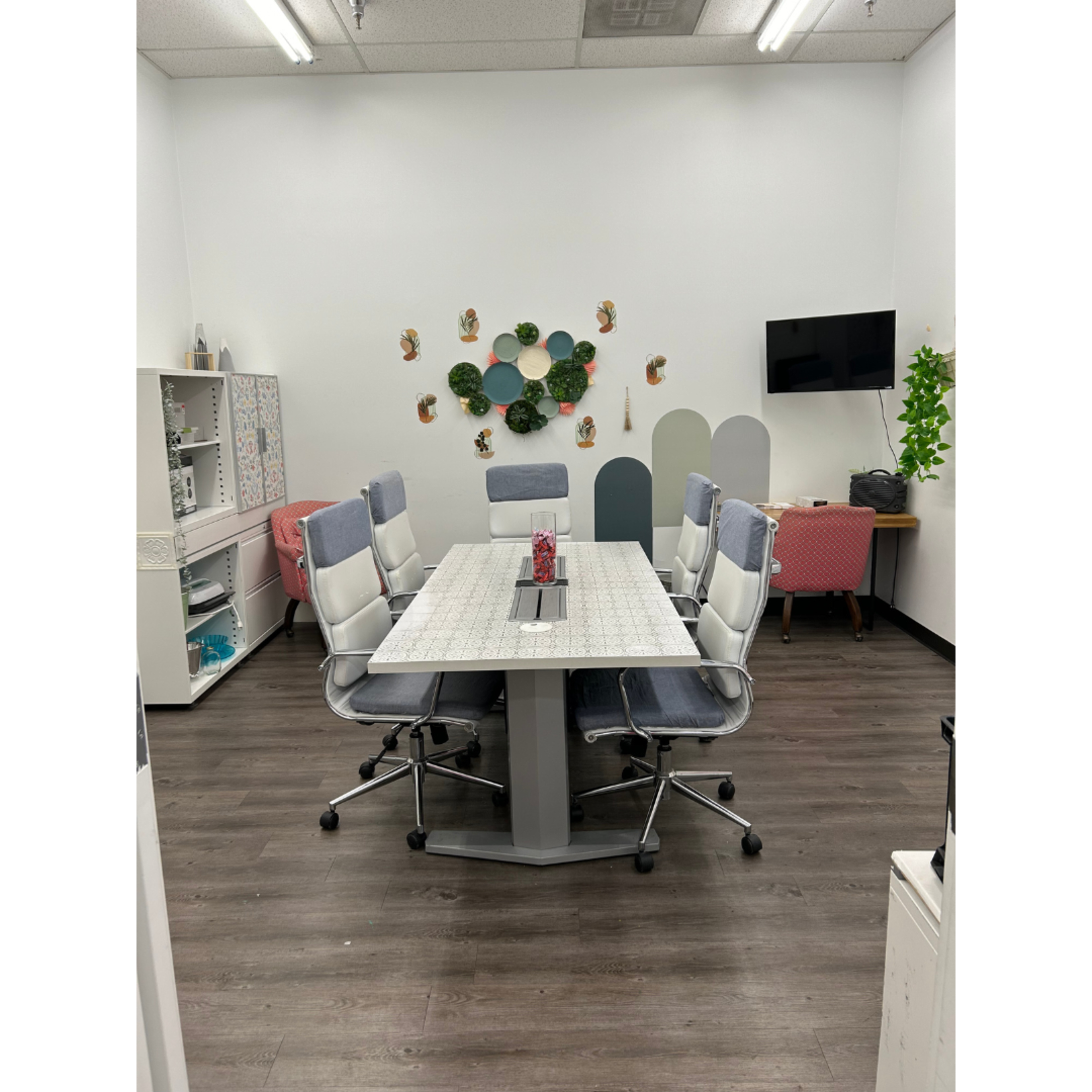 A modern meeting room featuring a large table with six ergonomic chairs, a decorative wall arrangement, and a television mounted on one wall.