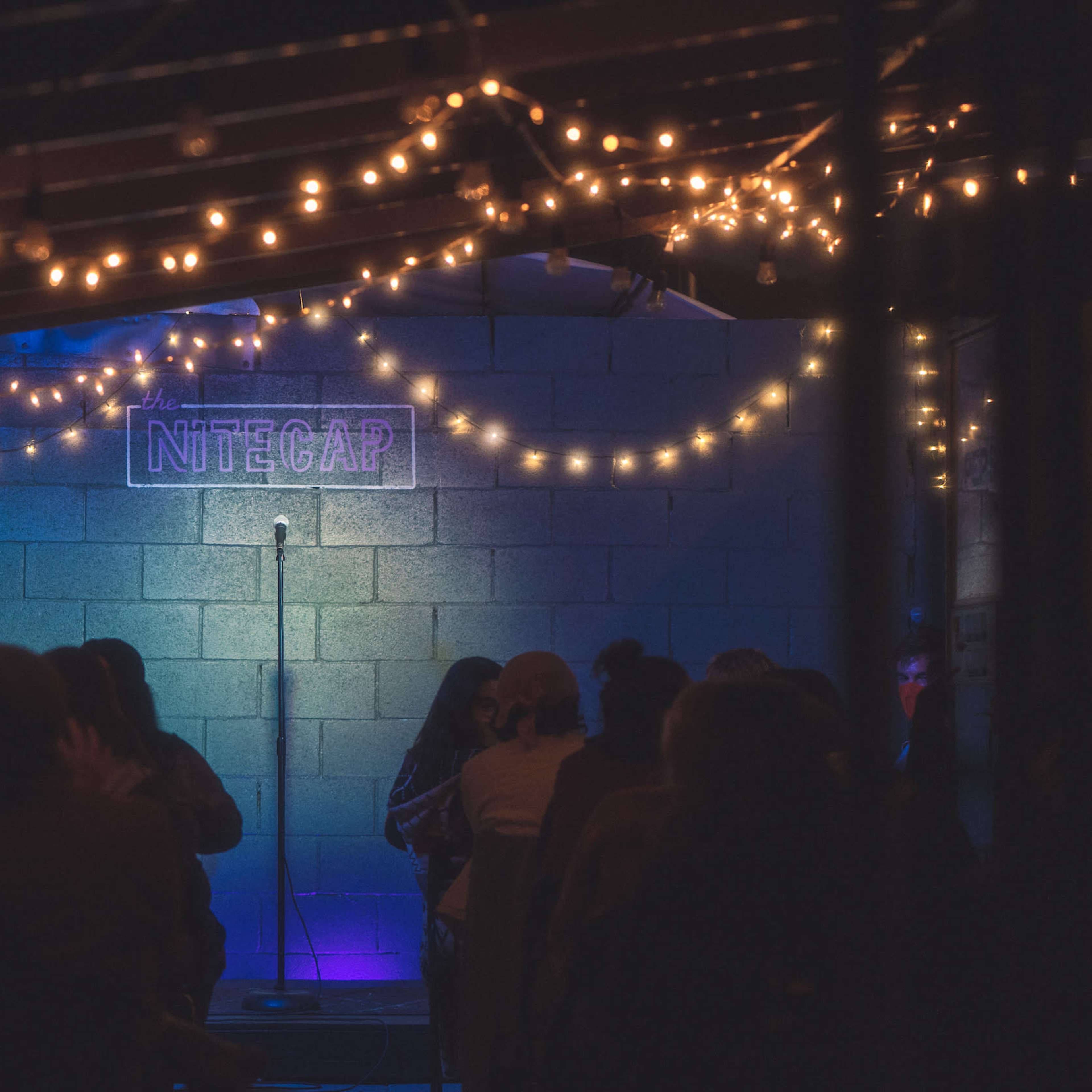 A dimly lit performance space features a microphone on stage, surrounded by string lights, with an audience seated in front.