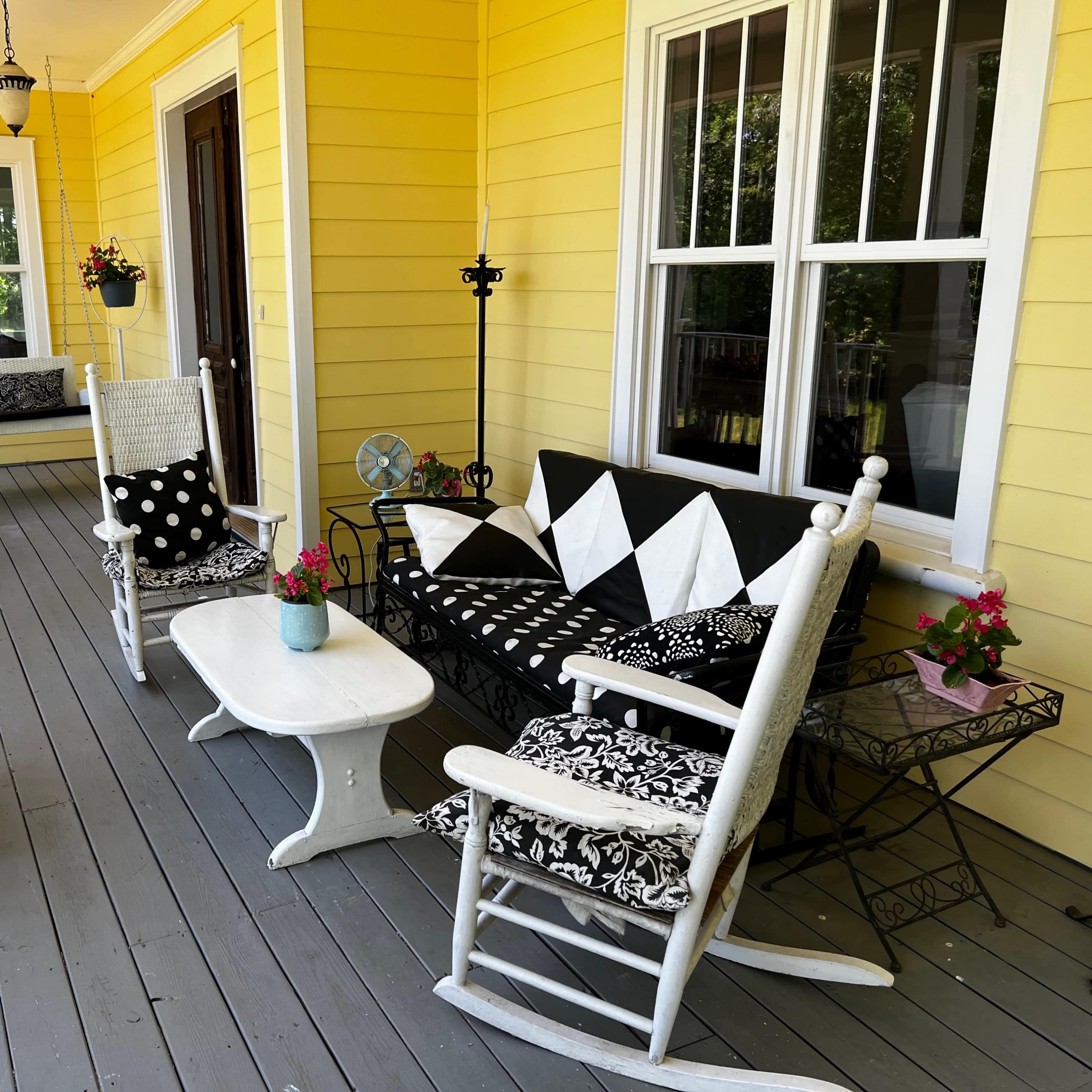 A bright yellow porch featuring white rocking chairs, a black and white upholstered bench, a small white table, and various potted flowers.