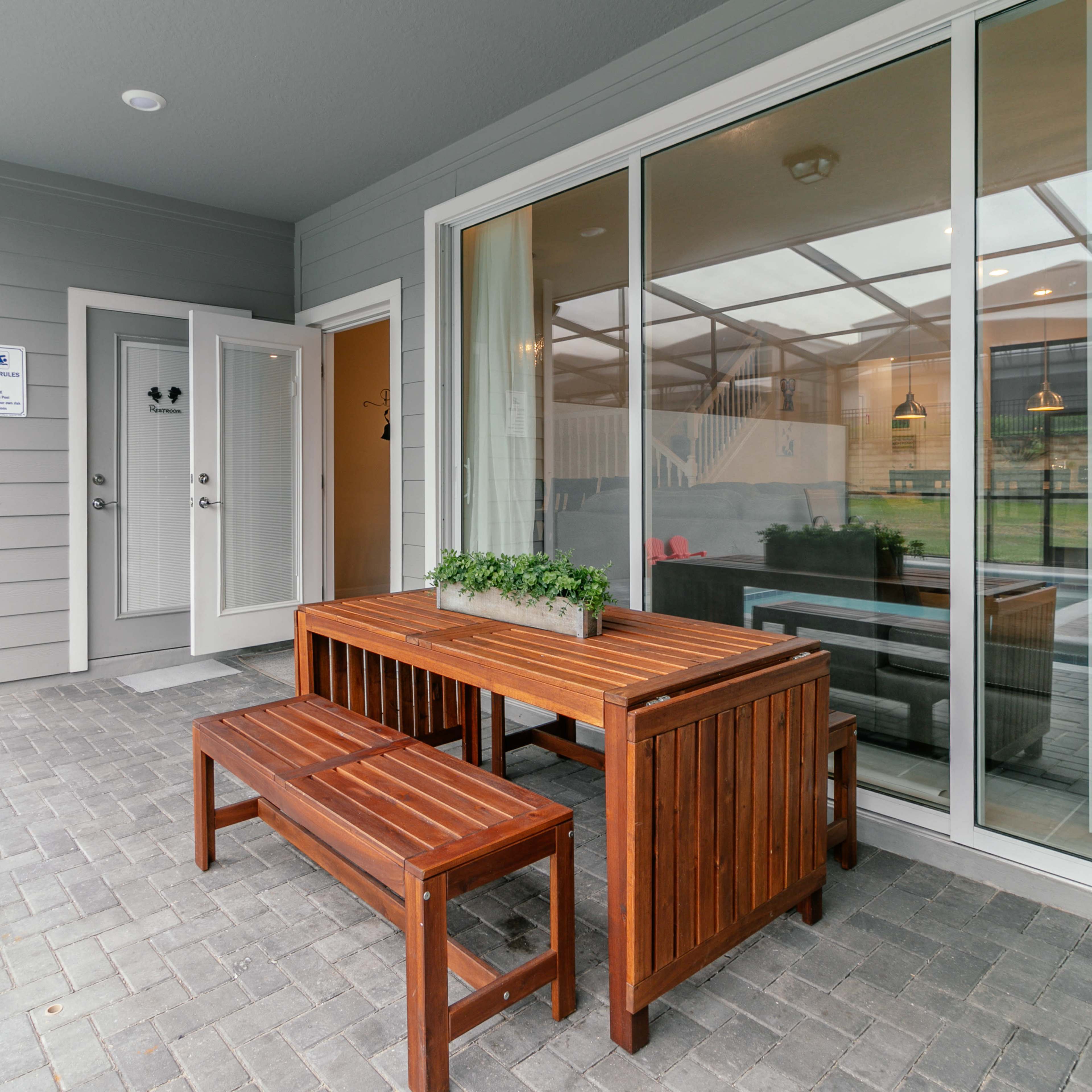A wooden table and bench set is placed on a brick patio outside a glass-enclosed room with a view of a lawn.