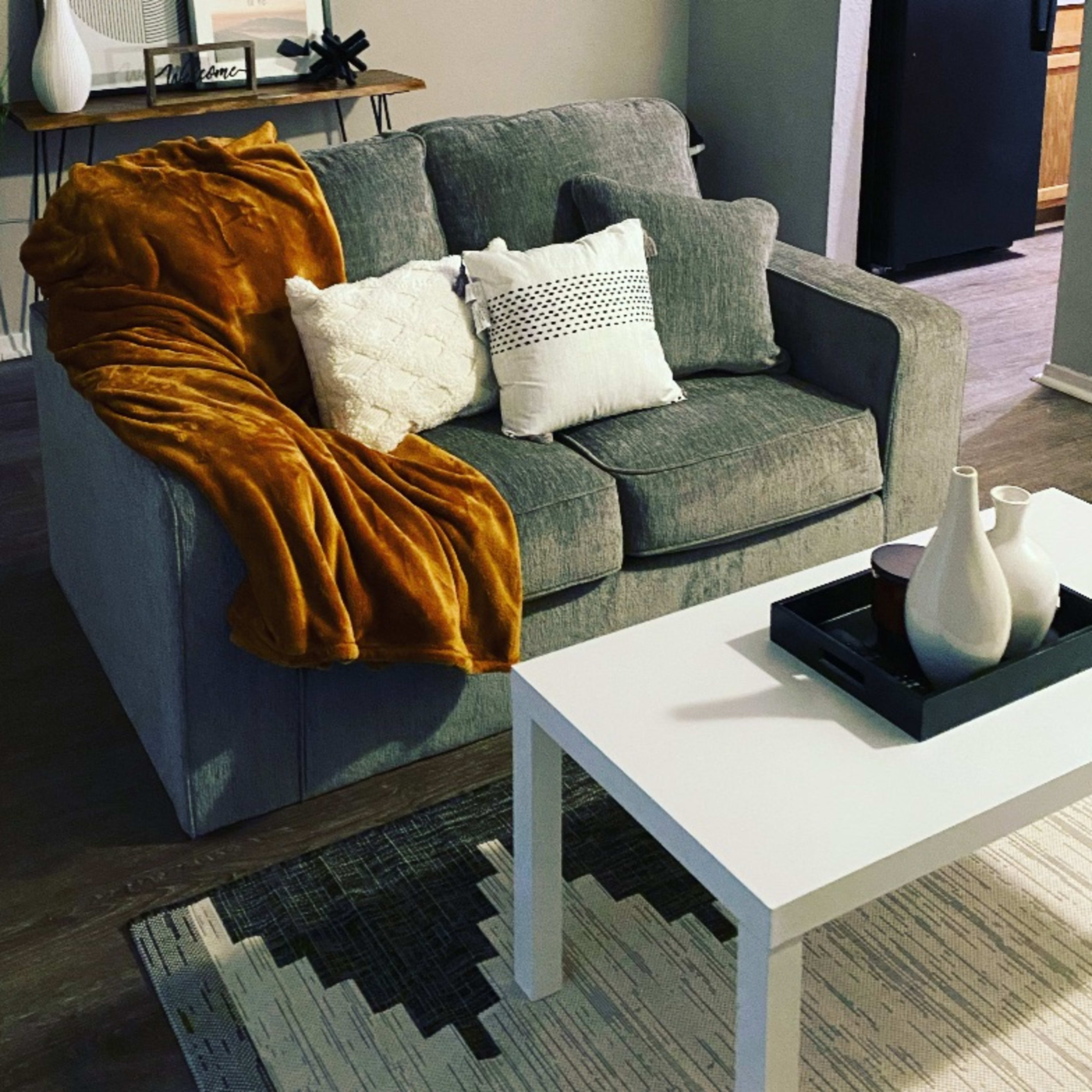 A gray couch with a mustard-colored throw blanket and several decorative pillows is positioned beside a white coffee table on a patterned rug in a living room.