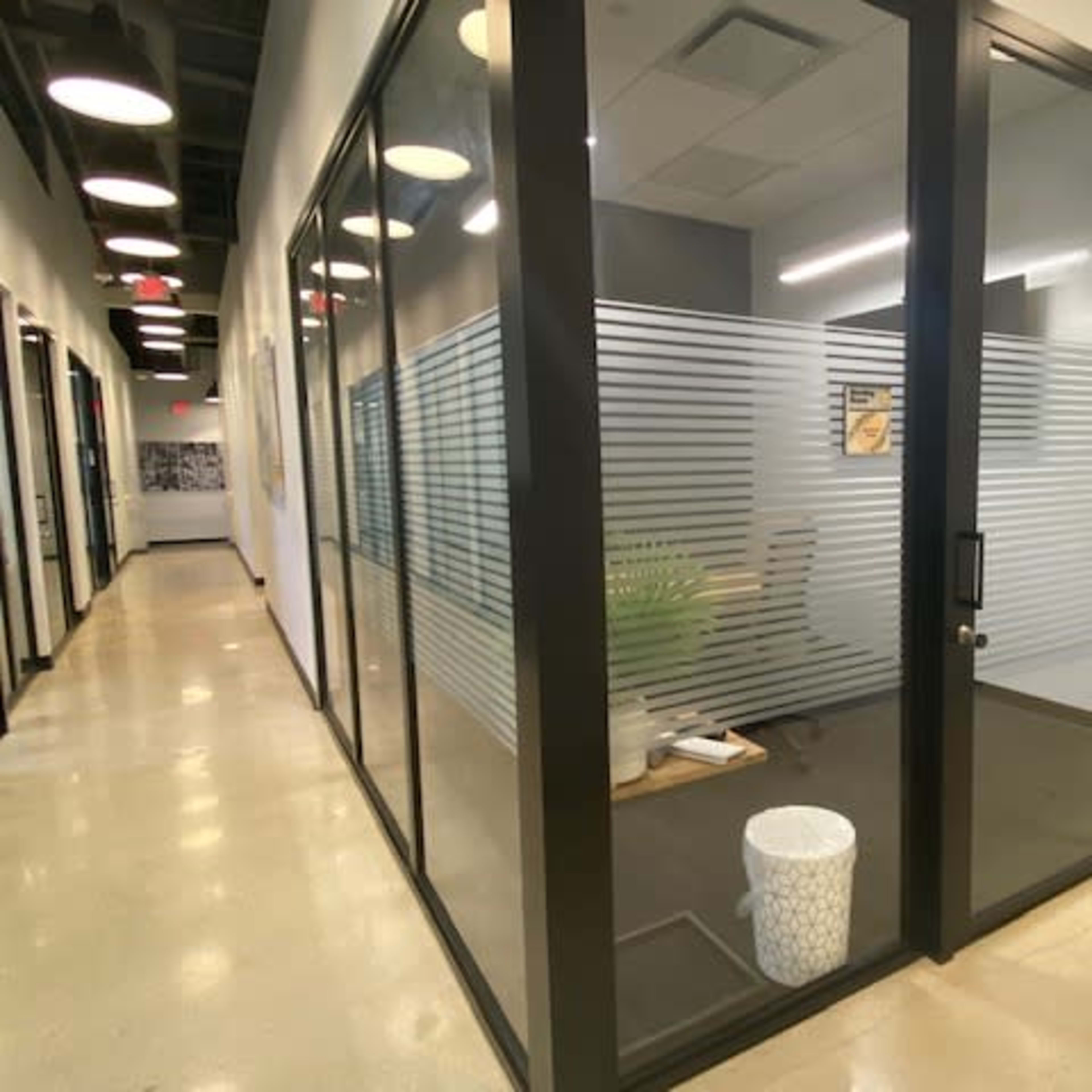 A modern glass-walled office with a small plant and a wastebasket, located in a corridor with overhead lighting.