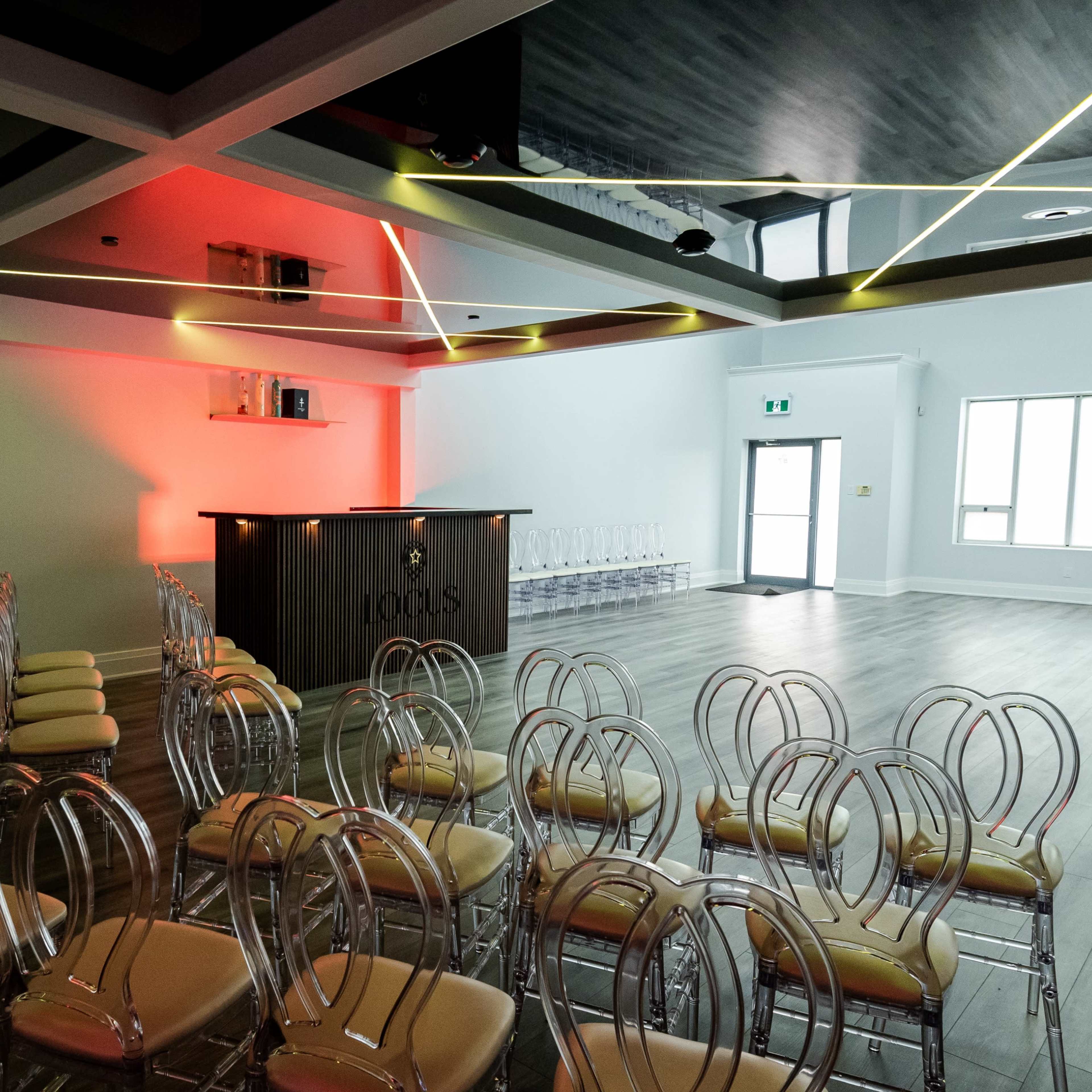 A conference room features rows of acrylic chairs facing a wooden bar with backlighting, set against a modern ceiling design.