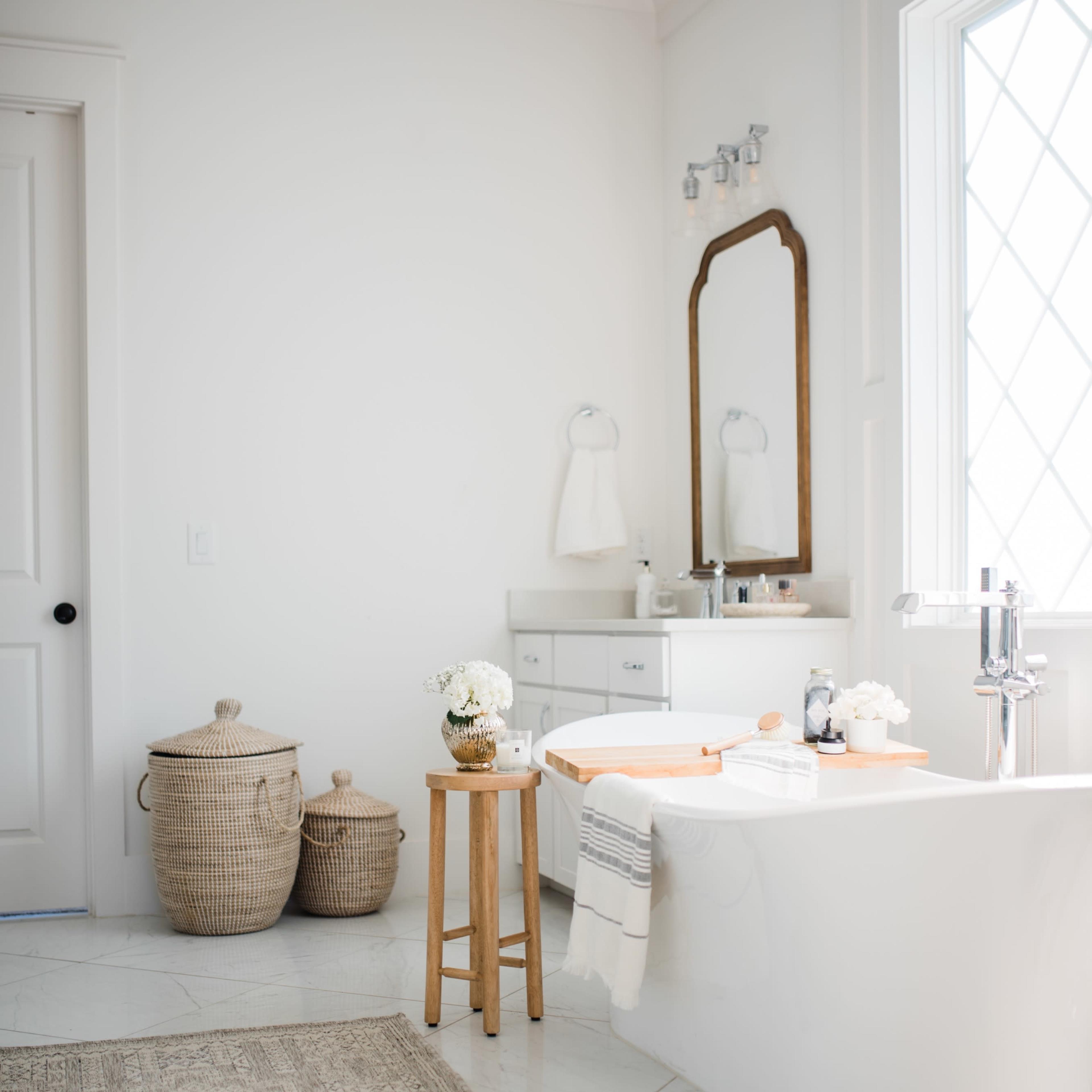 A neatly arranged bathroom features a freestanding bathtub, a wooden stool with decorative items, and two woven baskets beside a large window.
