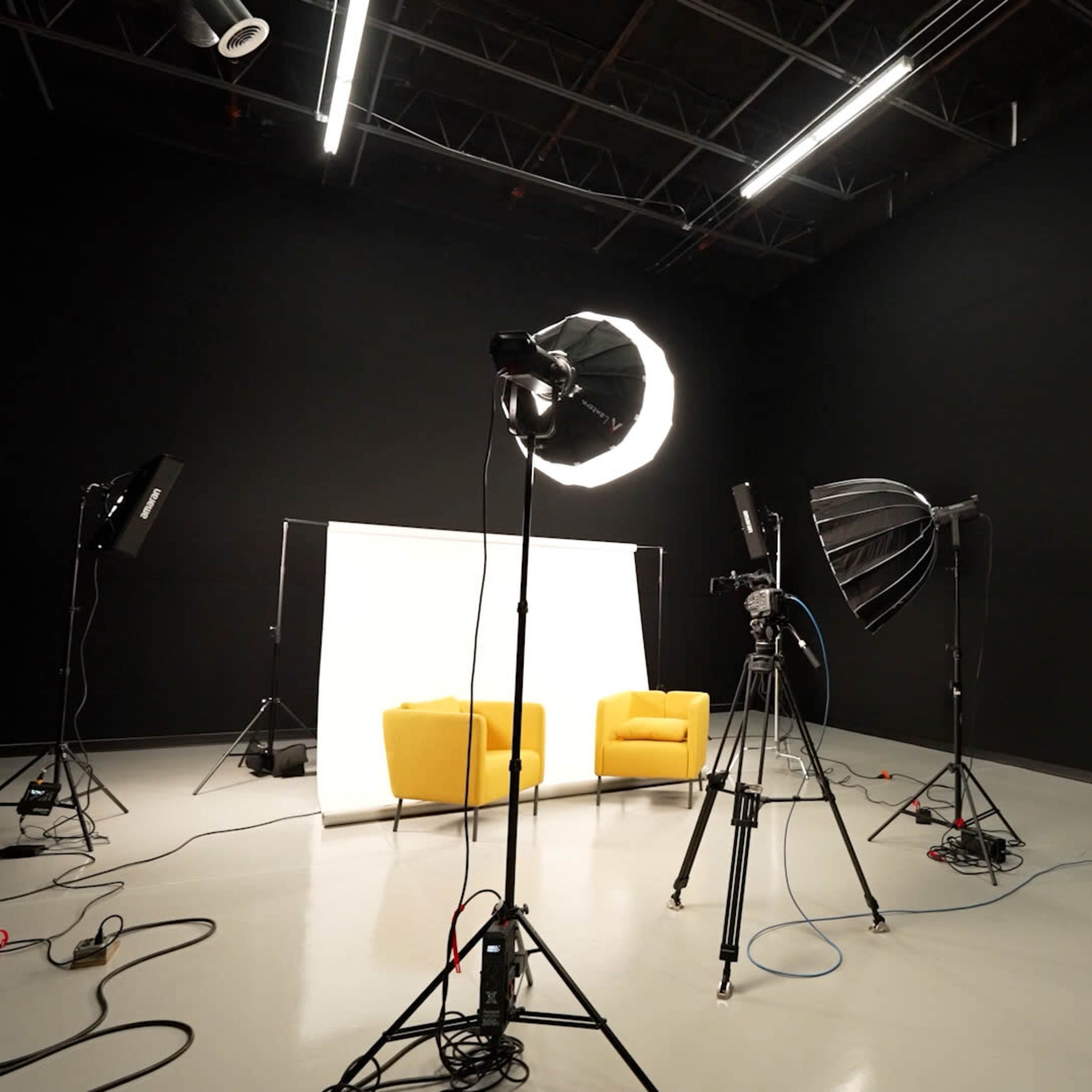 A dark studio space set up for a photoshoot, featuring two yellow chairs, lighting equipment, and a backdrop.