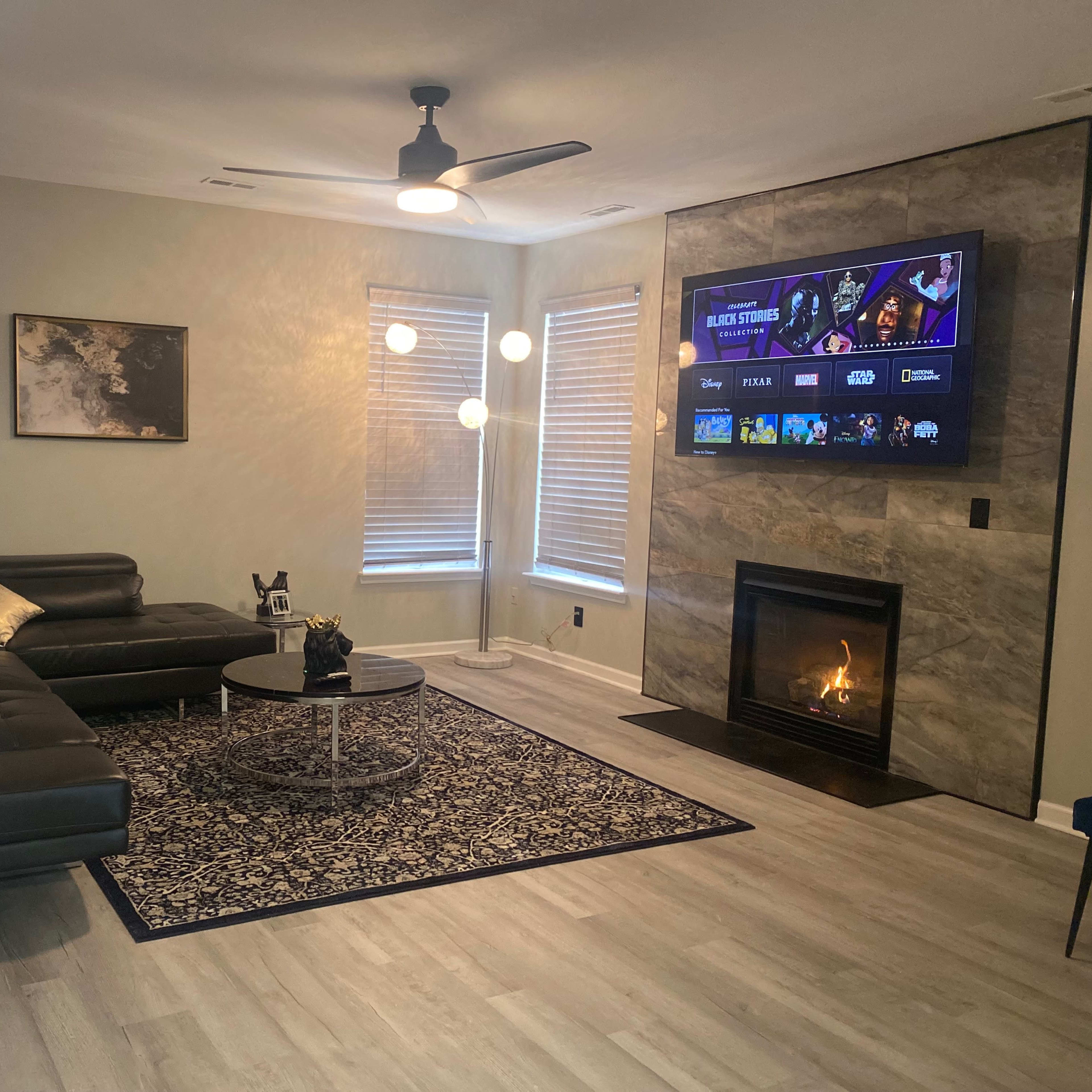 A modern living room with a sectional sofa, a circular coffee table, a large wall-mounted TV, and a fireplace beneath it, all arranged on light-colored flooring.