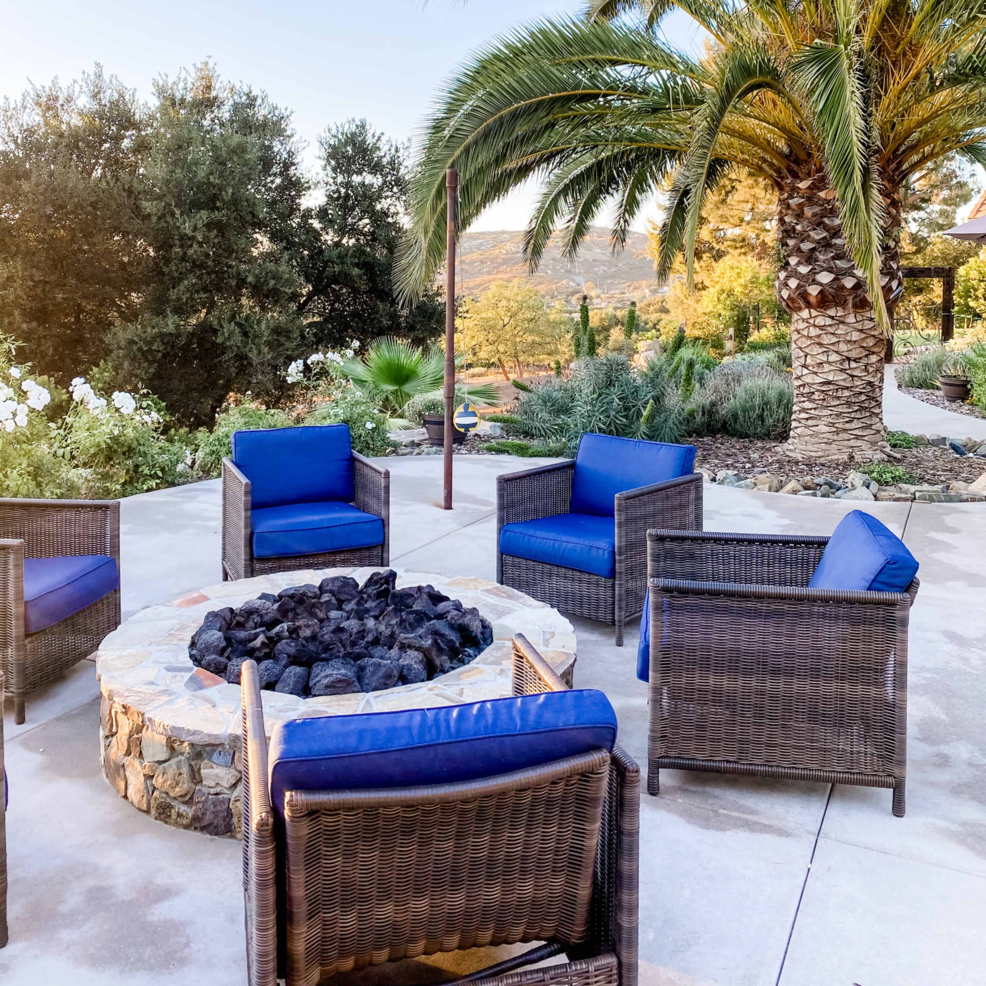 A circular stone fire pit is surrounded by blue cushioned chairs under a palm tree and overlooking a landscaped area.