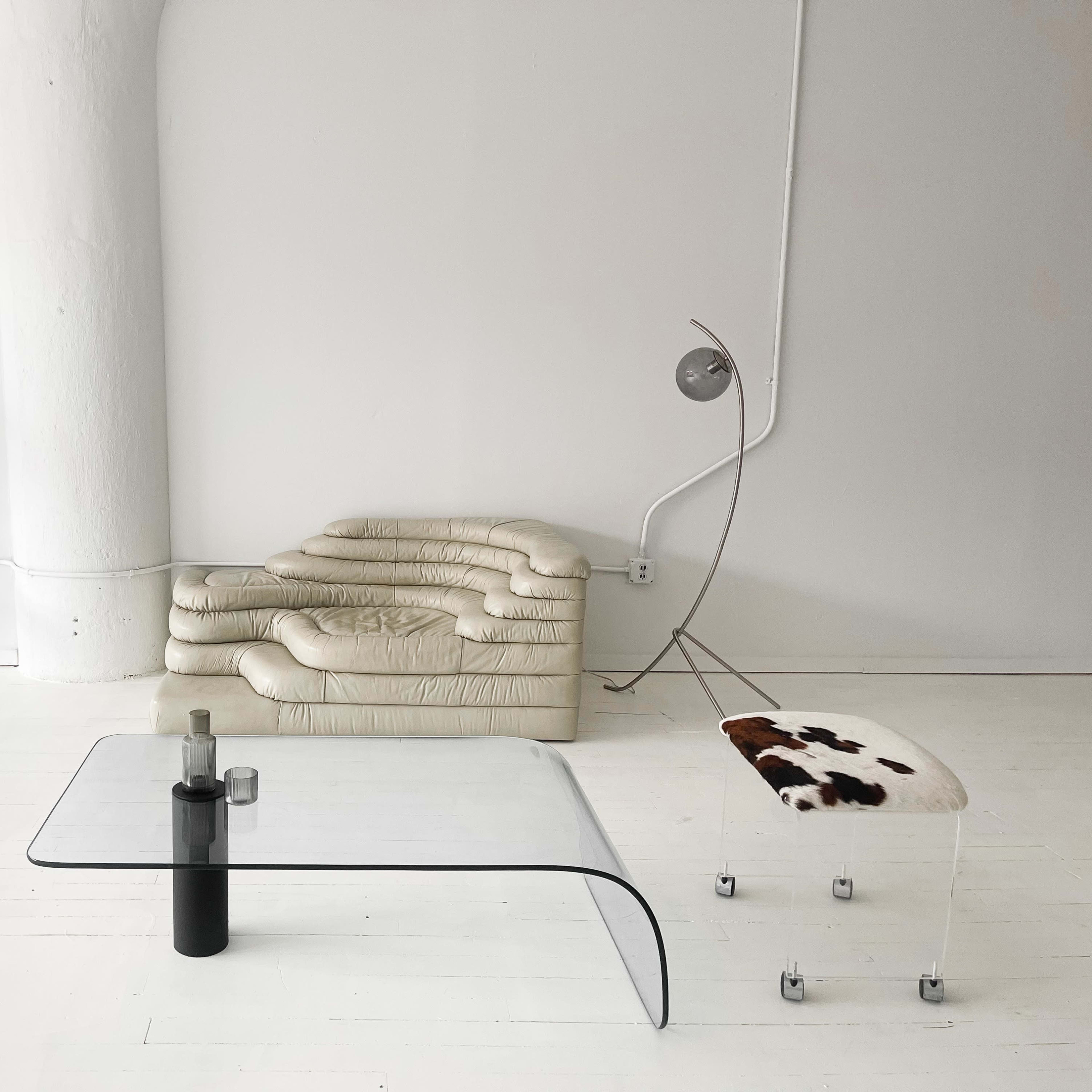 A minimalist interior with a clear glass coffee table, a curved beige sofa, a transparent chair, and a unique bench with a cow print design.
