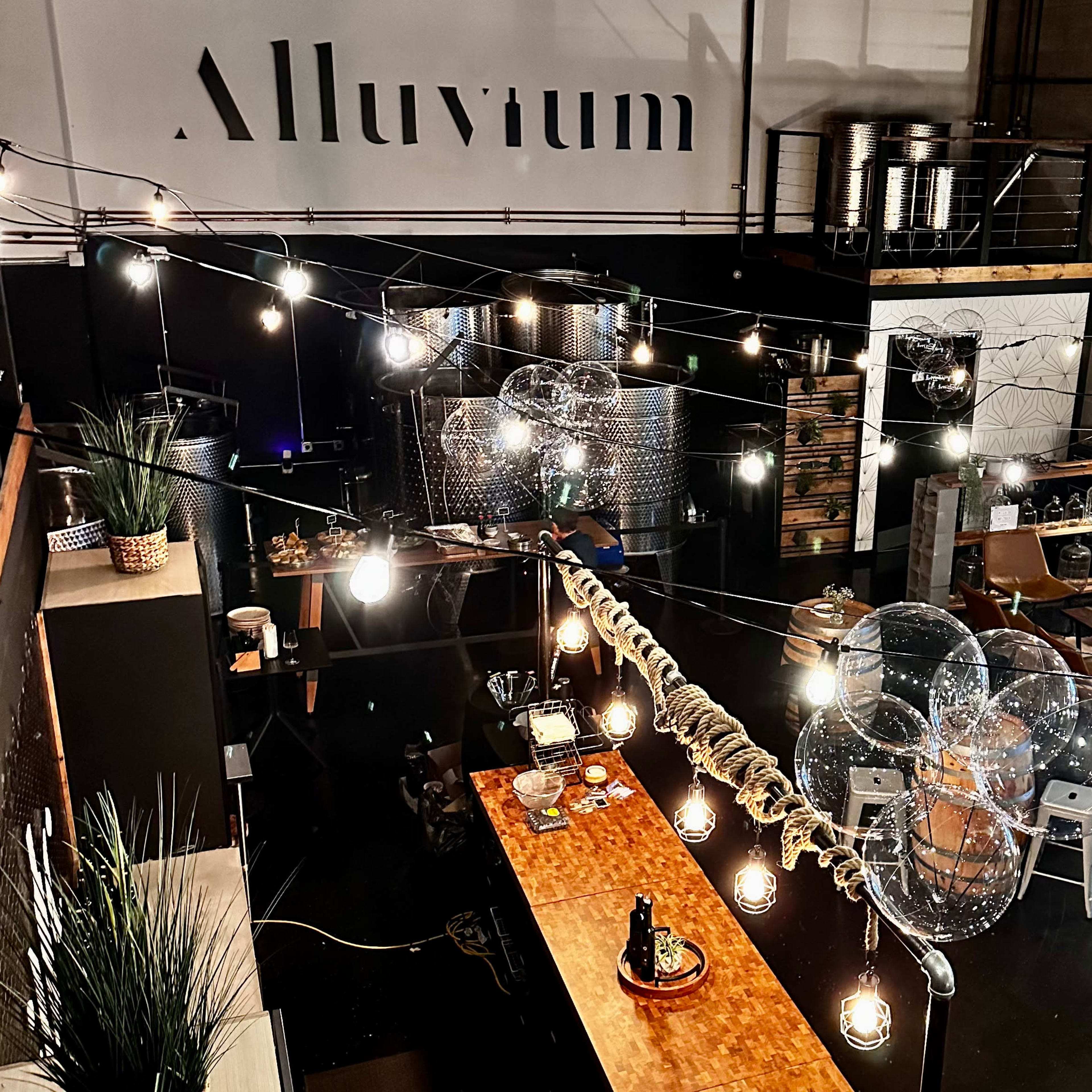 A spacious industrial-style room features a long wooden table adorned with plants and lights, surrounded by modern seating and large storage tanks in the background.