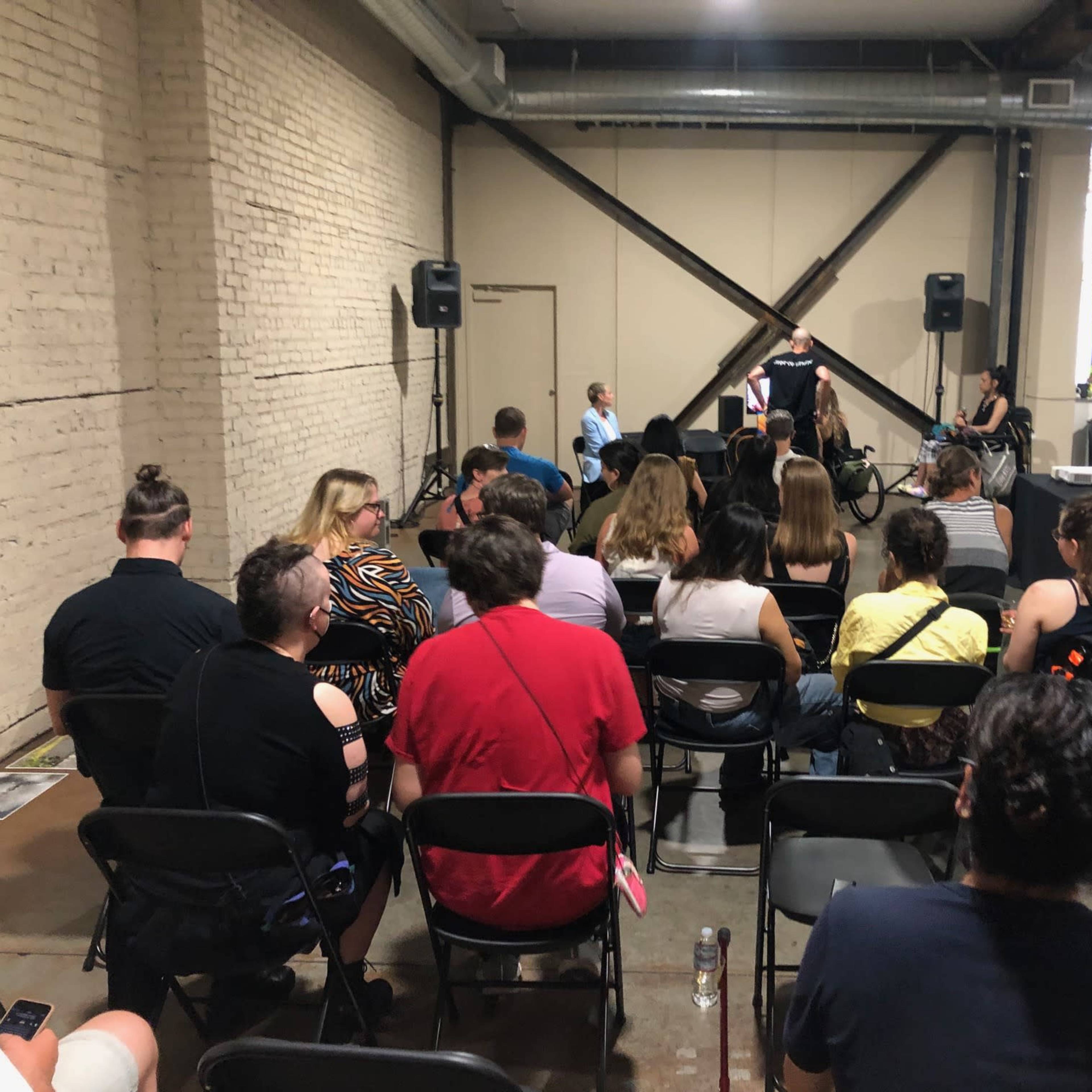 A group of people sits in rows of chairs facing a small stage in a sparsely decorated indoor space.
