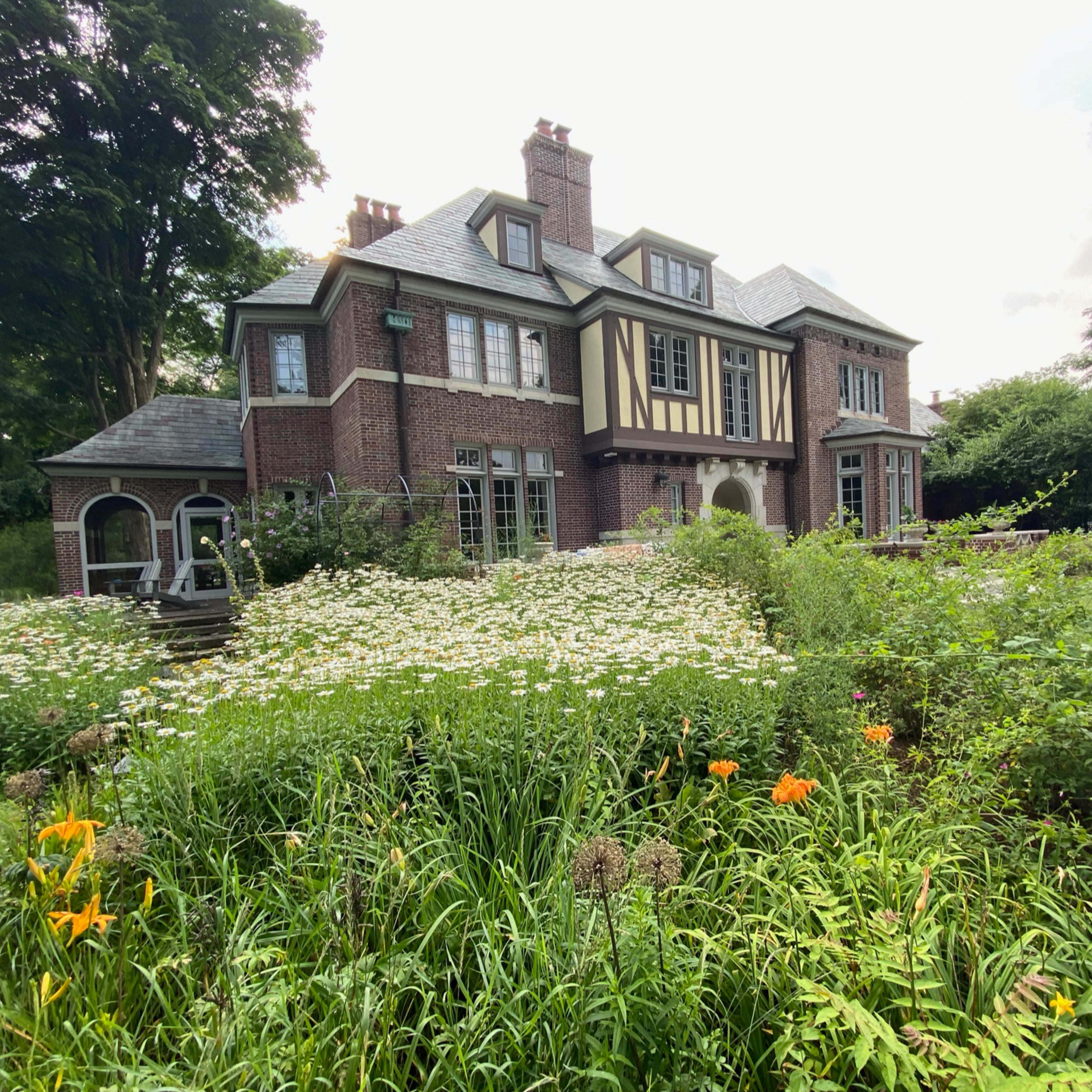 A large, brick house with a gabled roof is surrounded by a garden filled with various flowers and greenery.