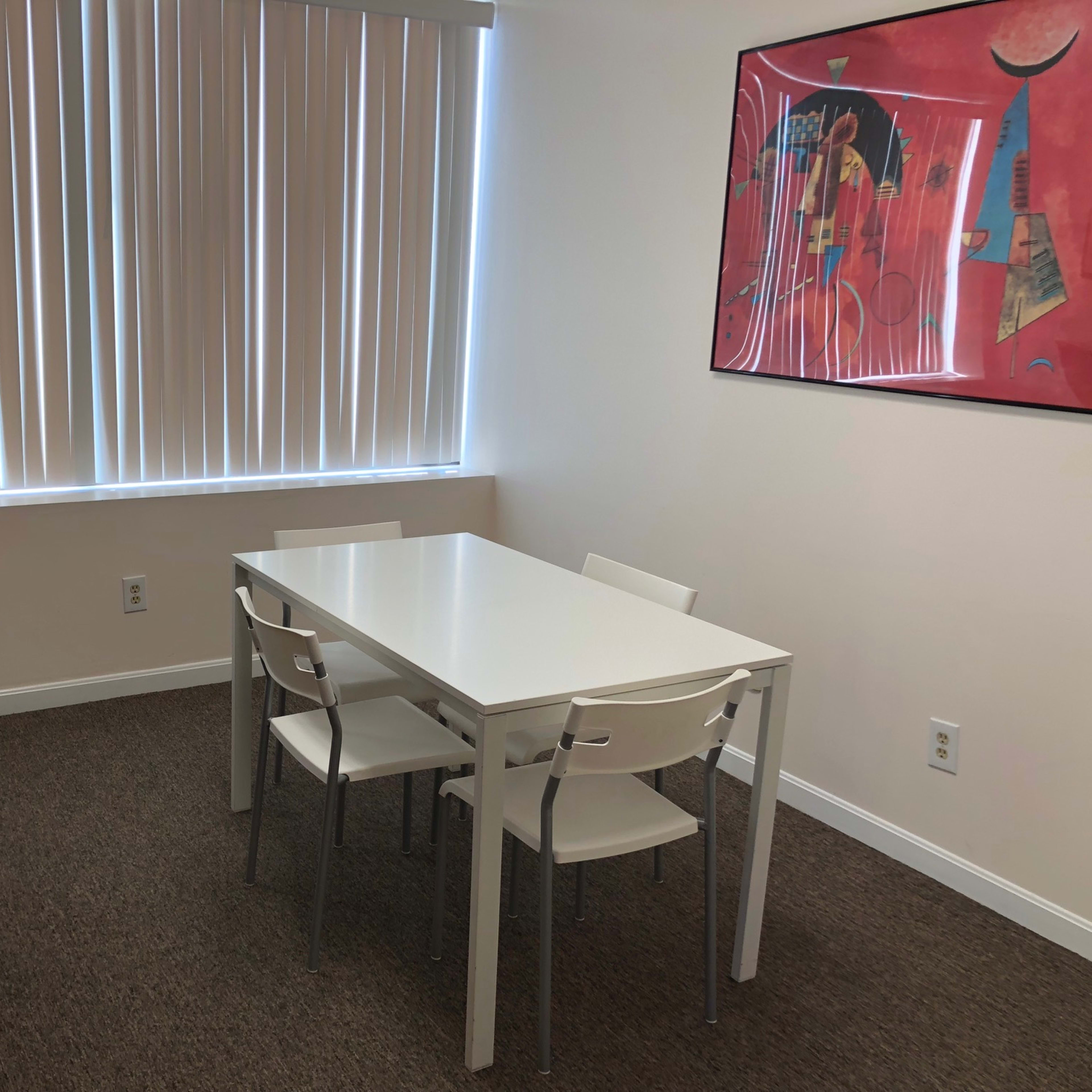 A white table with four chairs is set against a wall with a window and a colorful abstract painting, next to a brown piano.