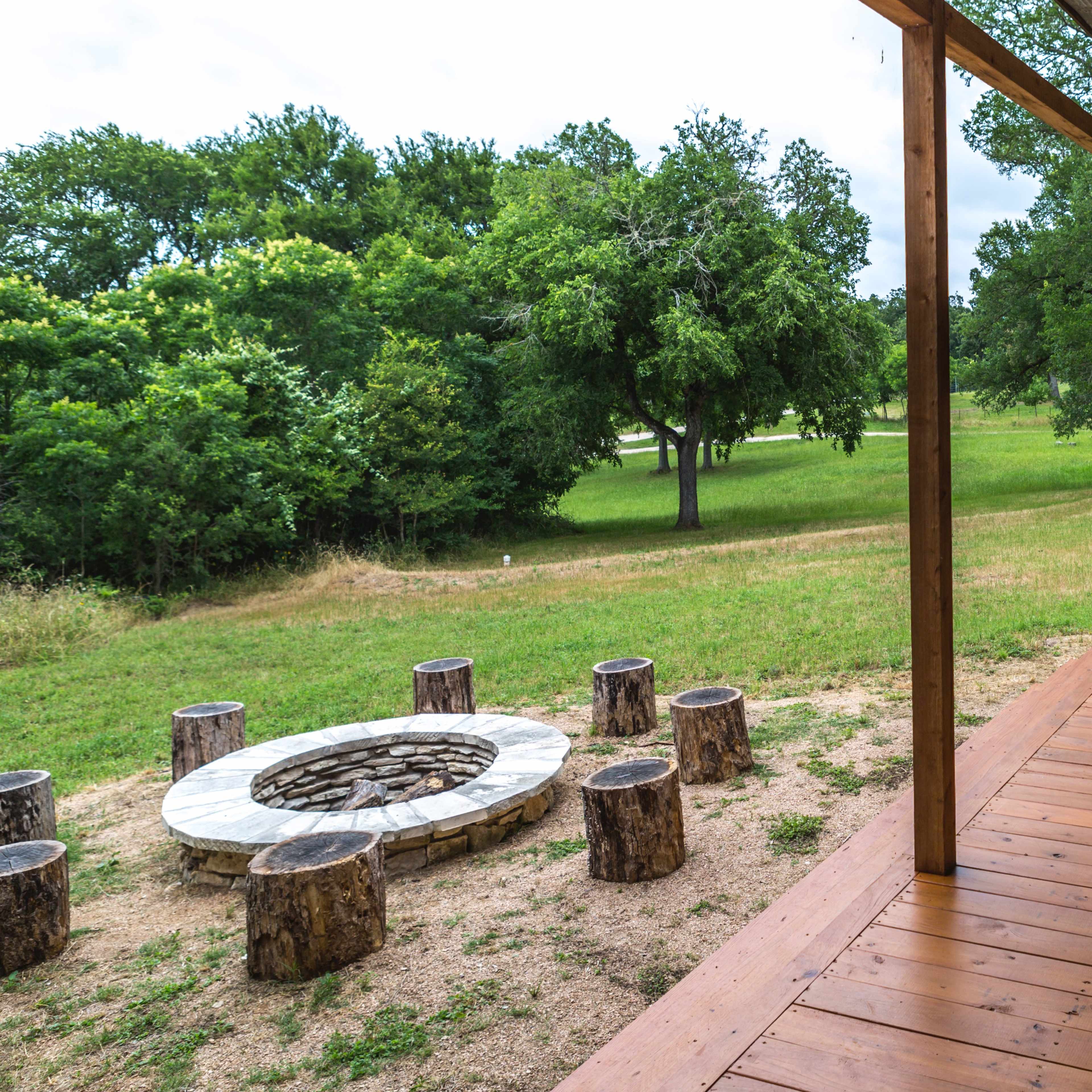 A wooden porch overlooks a stone fire pit surrounded by log seating, set against a backdrop of green trees and open grass.