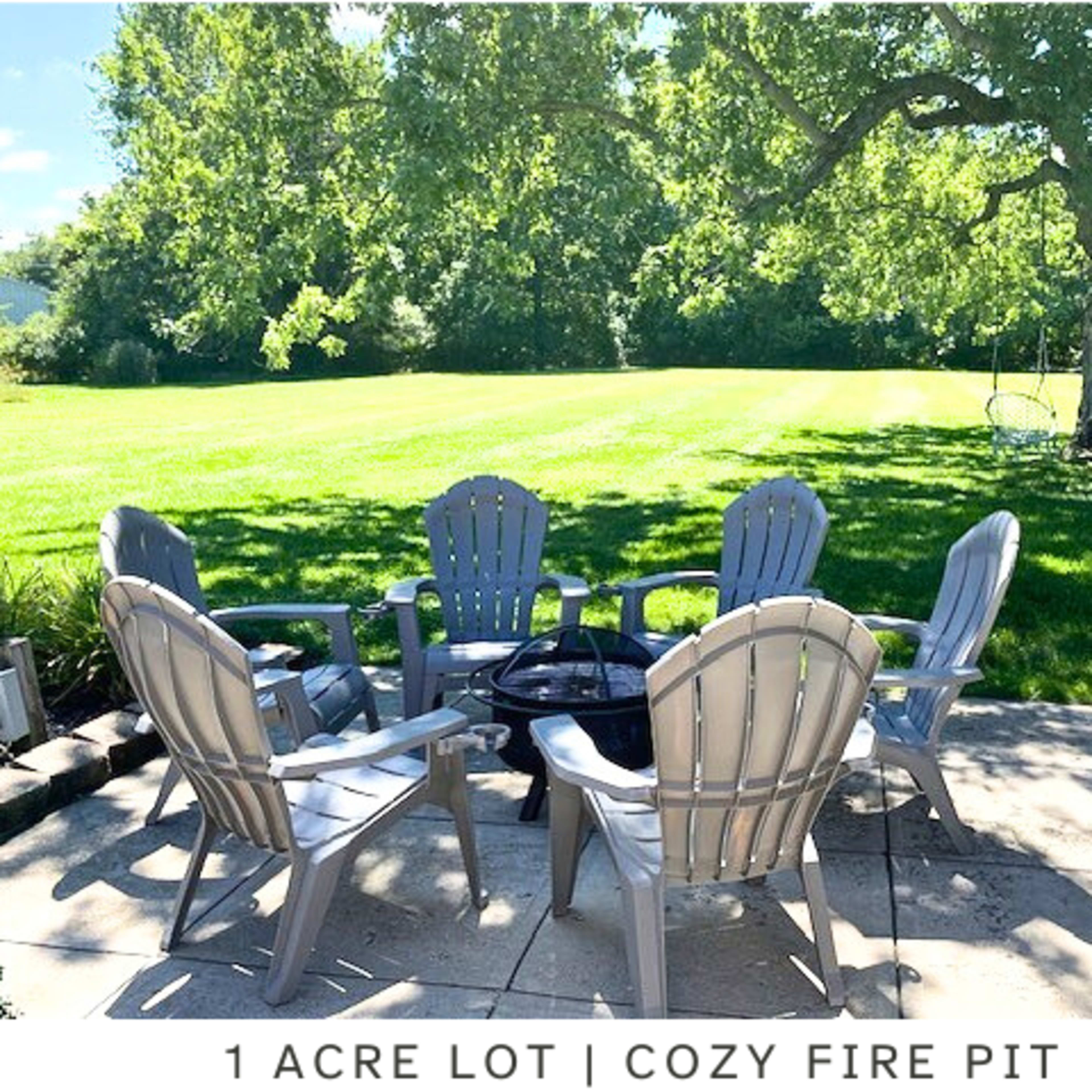 A spacious backyard with a circular arrangement of six gray chairs around a fire pit on a stone patio, set in a grassy one-acre lot.