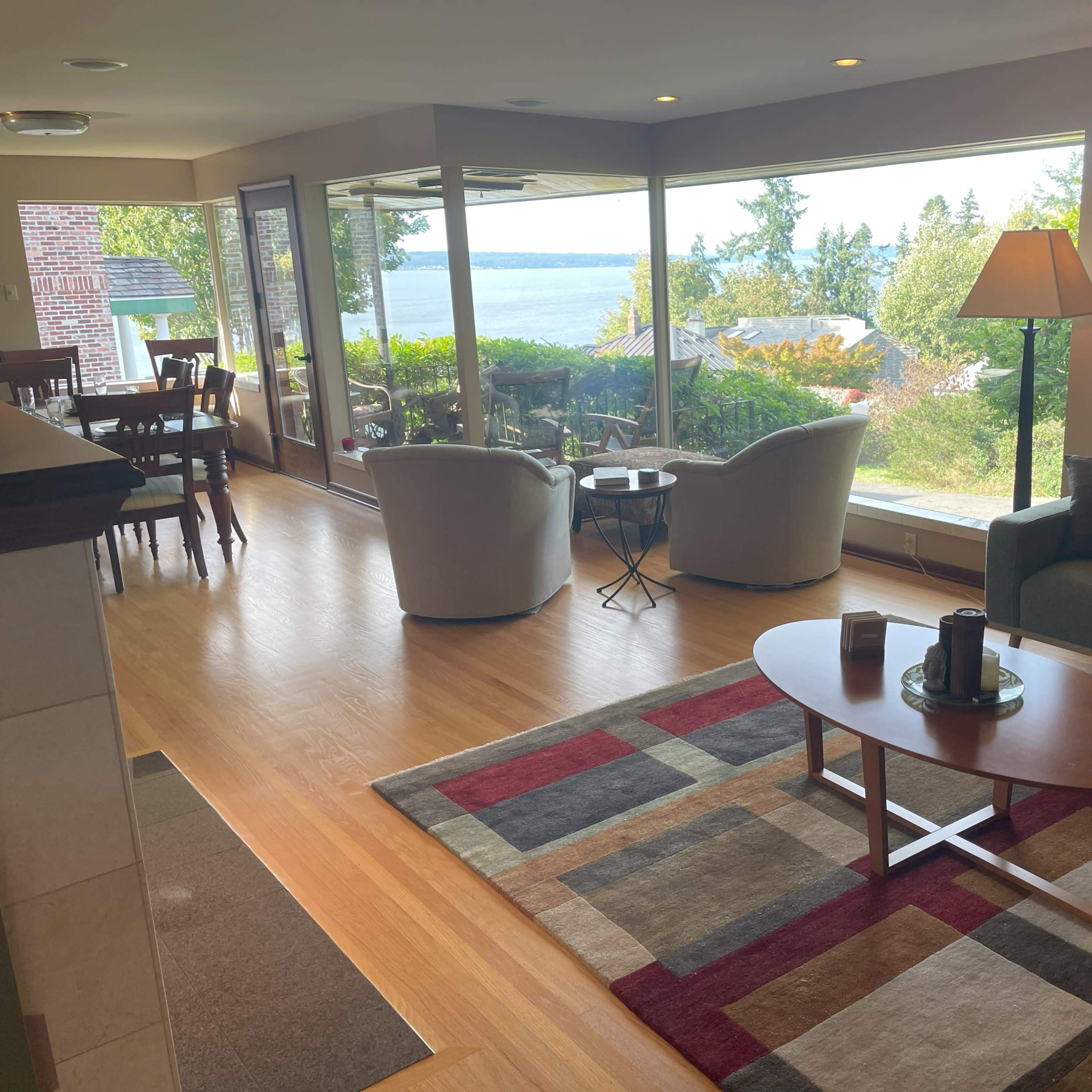 The living room features a couch, chairs, and a coffee table, with large windows overlooking a scenic view of trees and water.