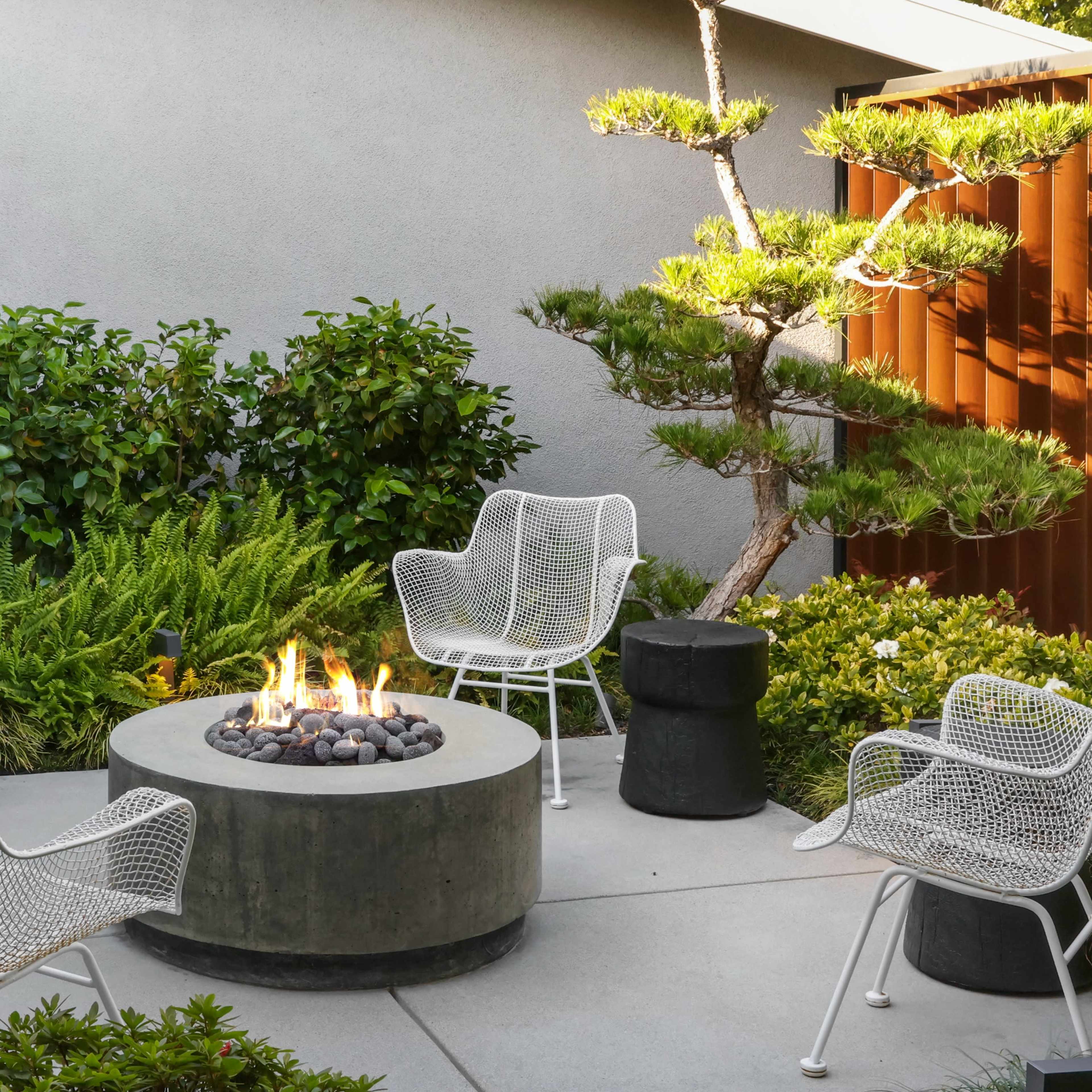 A circular concrete fire pit surrounded by four white wire chairs and black stool-like tables in a landscaped courtyard with greenery.