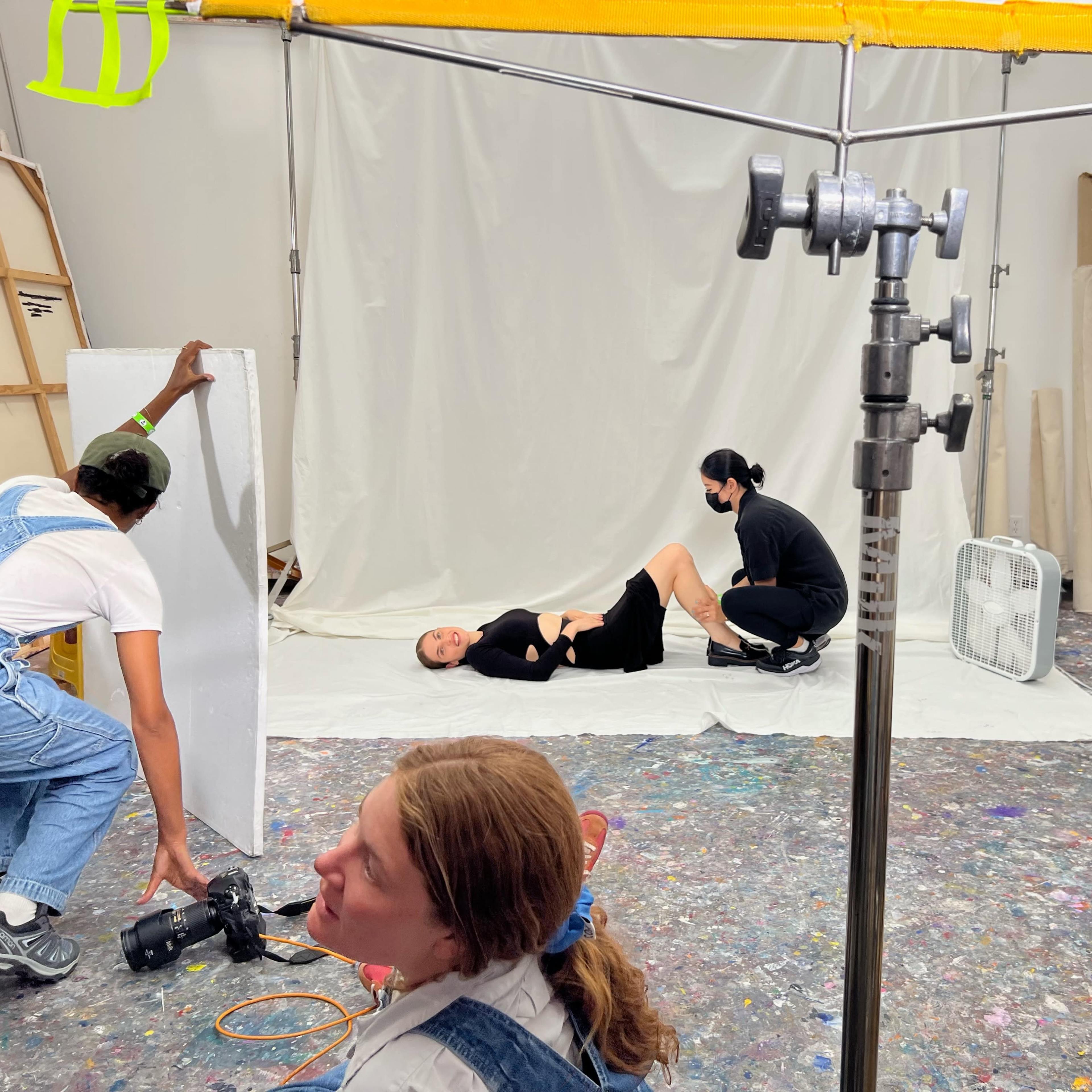 A photo shoot is taking place in a studio, with a person lying on a white backdrop while two people are positioned nearby, one adjusting equipment and another preparing to take a photo.