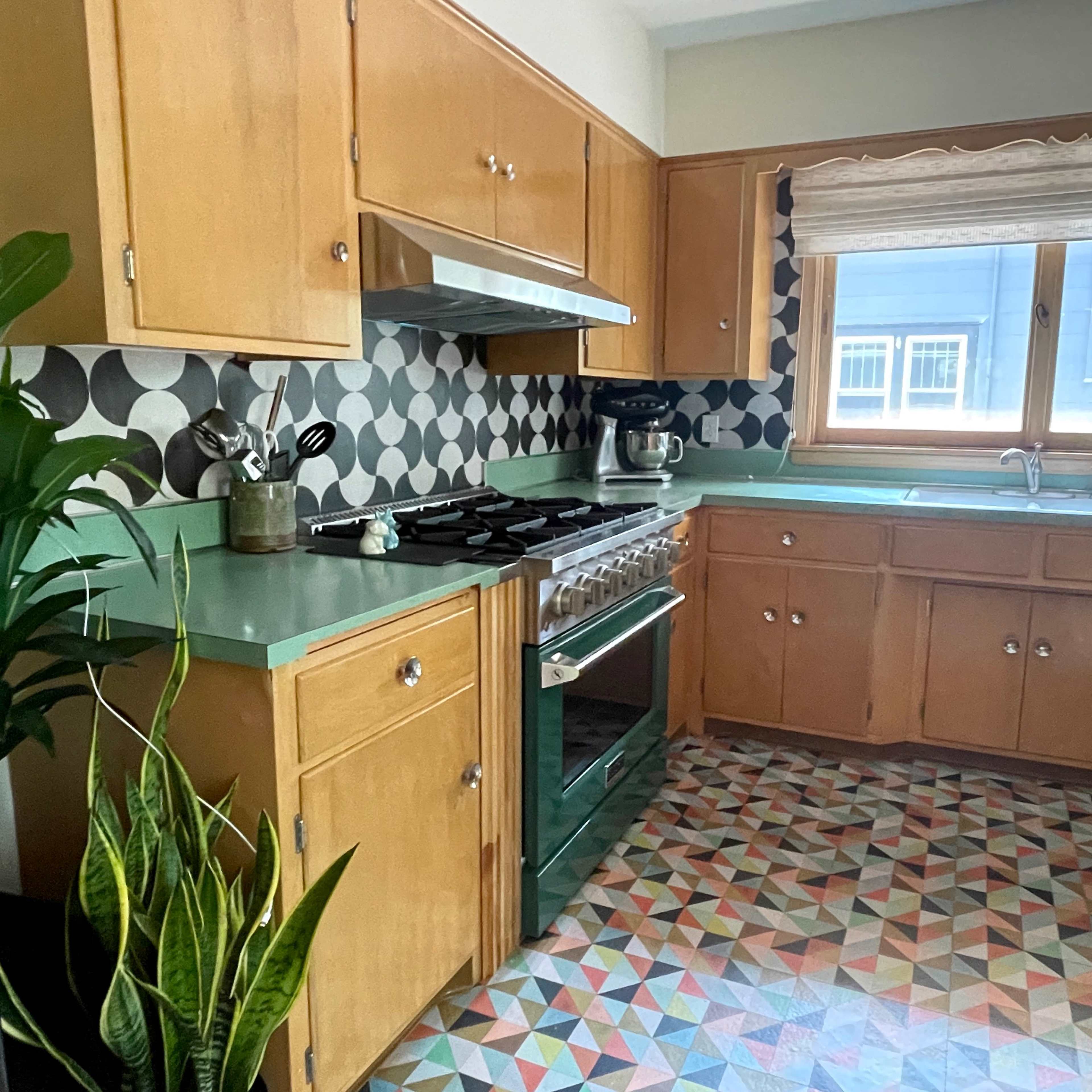 A kitchen features wooden cabinets, a stainless steel stove, and patterned tile flooring with a large window overlooking the outdoors.