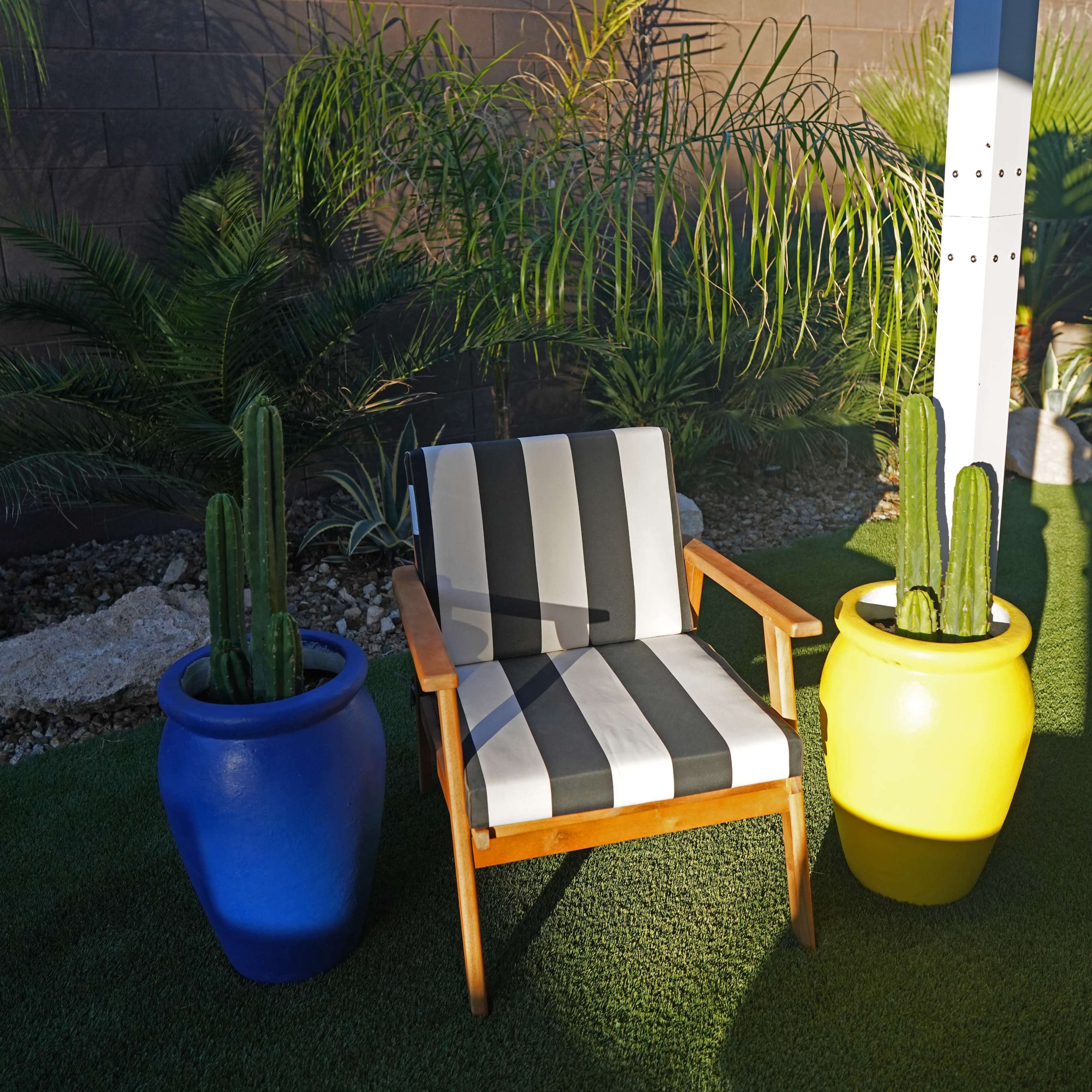 A striped chair sits between two potted cacti in a backyard with artificial grass and palm plants in the background.