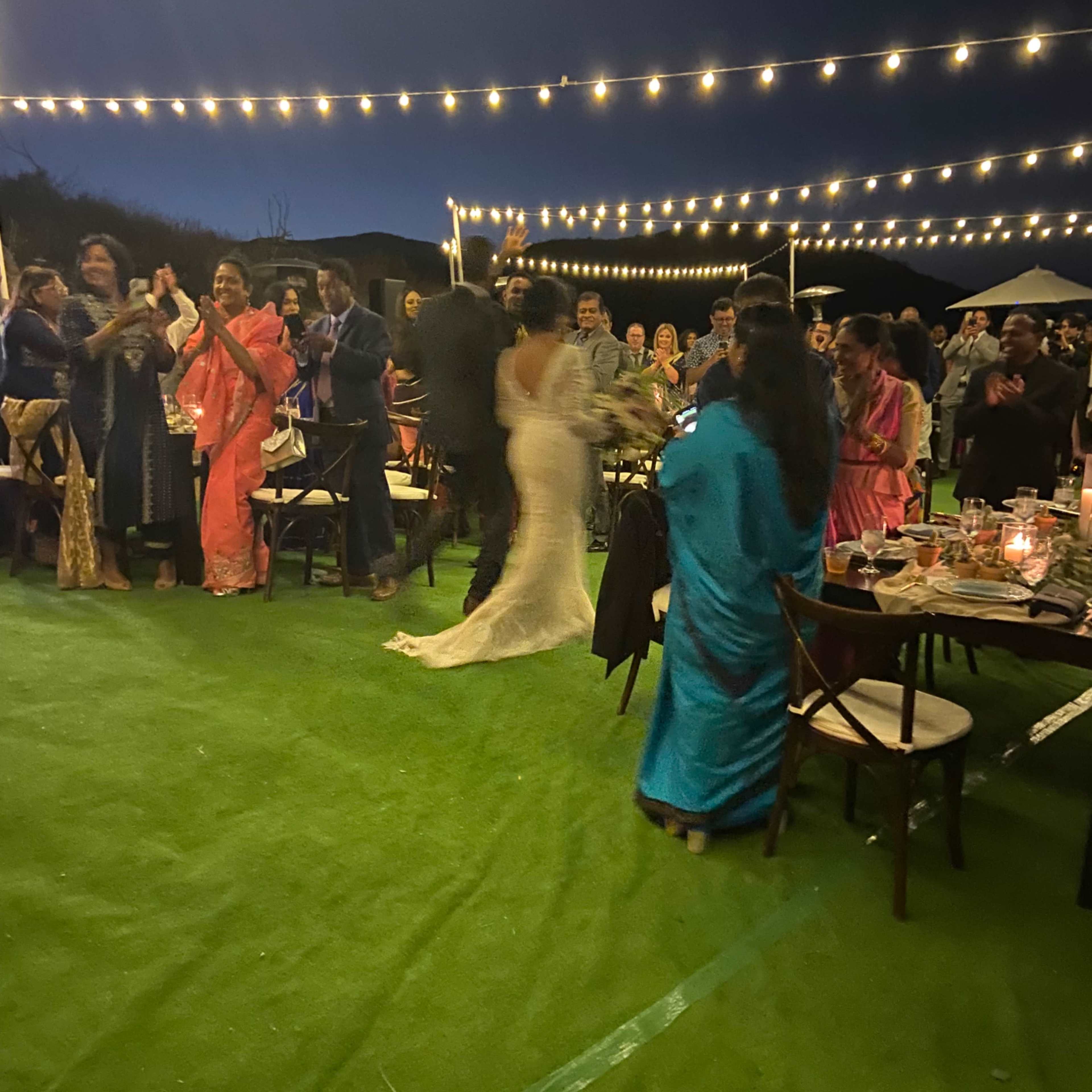 A large outdoor celebration is taking place at night with guests dancing and socializing under string lights.