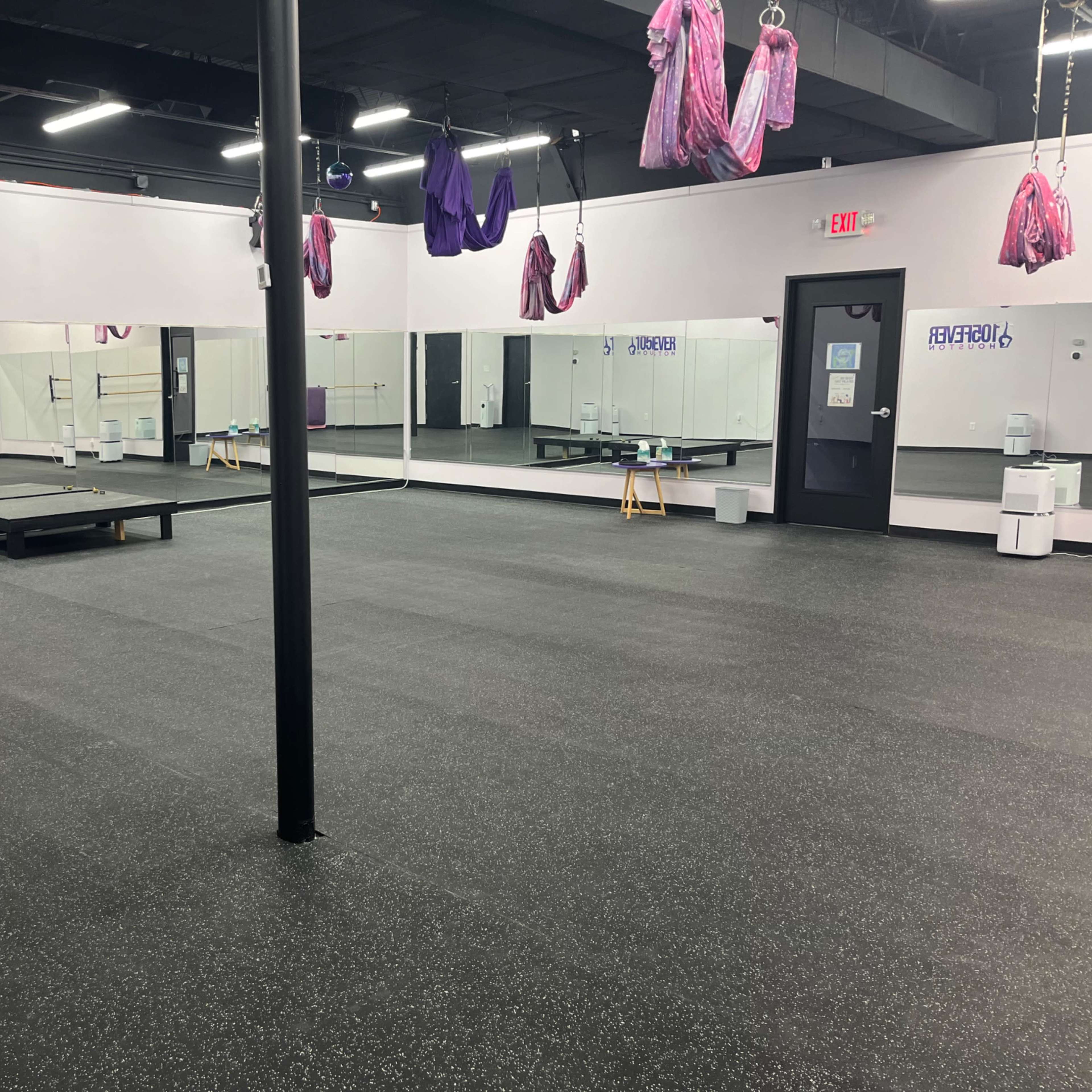 An empty fitness studio with a black rubber floor, a mirrored wall, and hanging exercise equipment from the ceiling.
