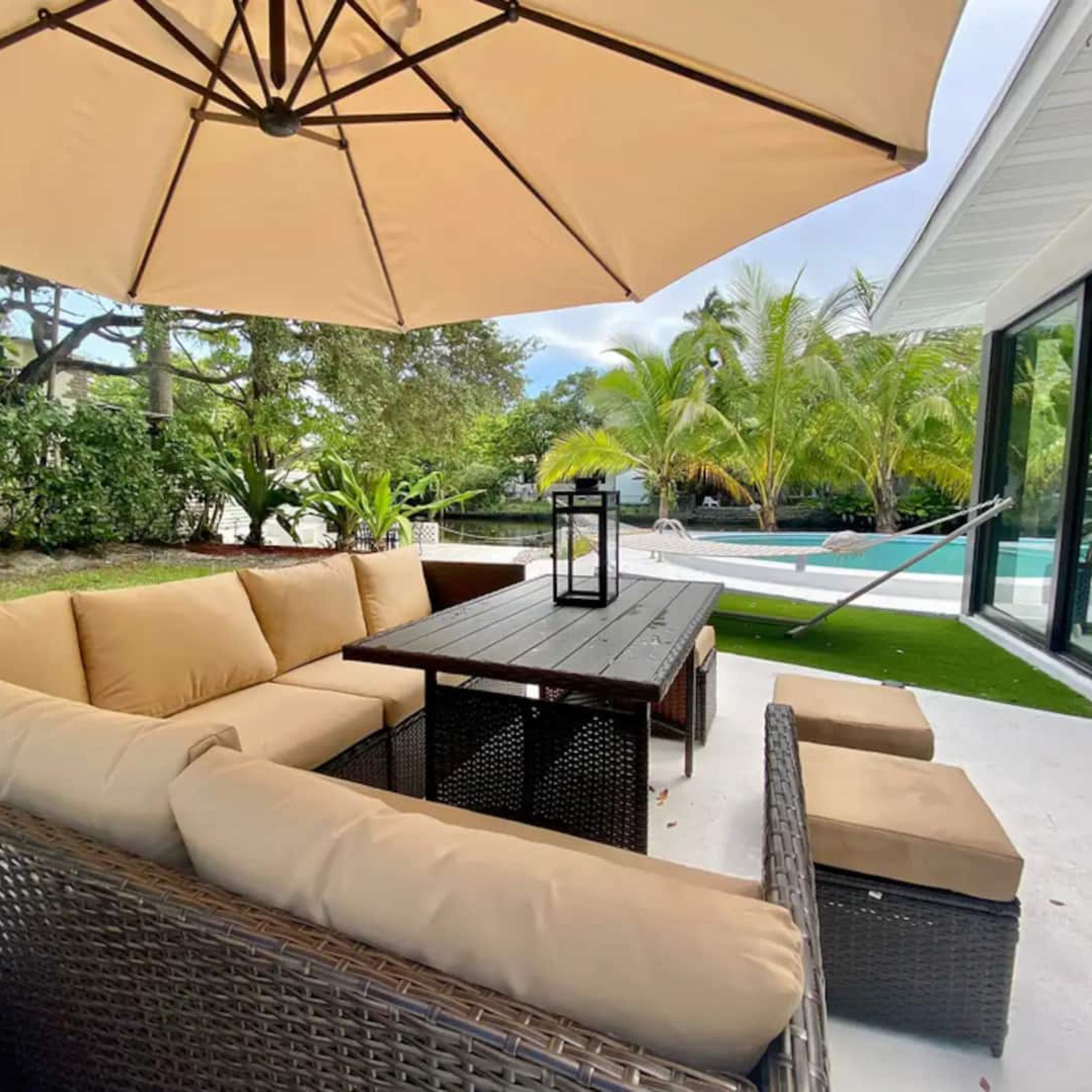 A patio area features a large sectional sofa underneath a beige umbrella, with a coffee table and a view of a pool surrounded by palm trees.