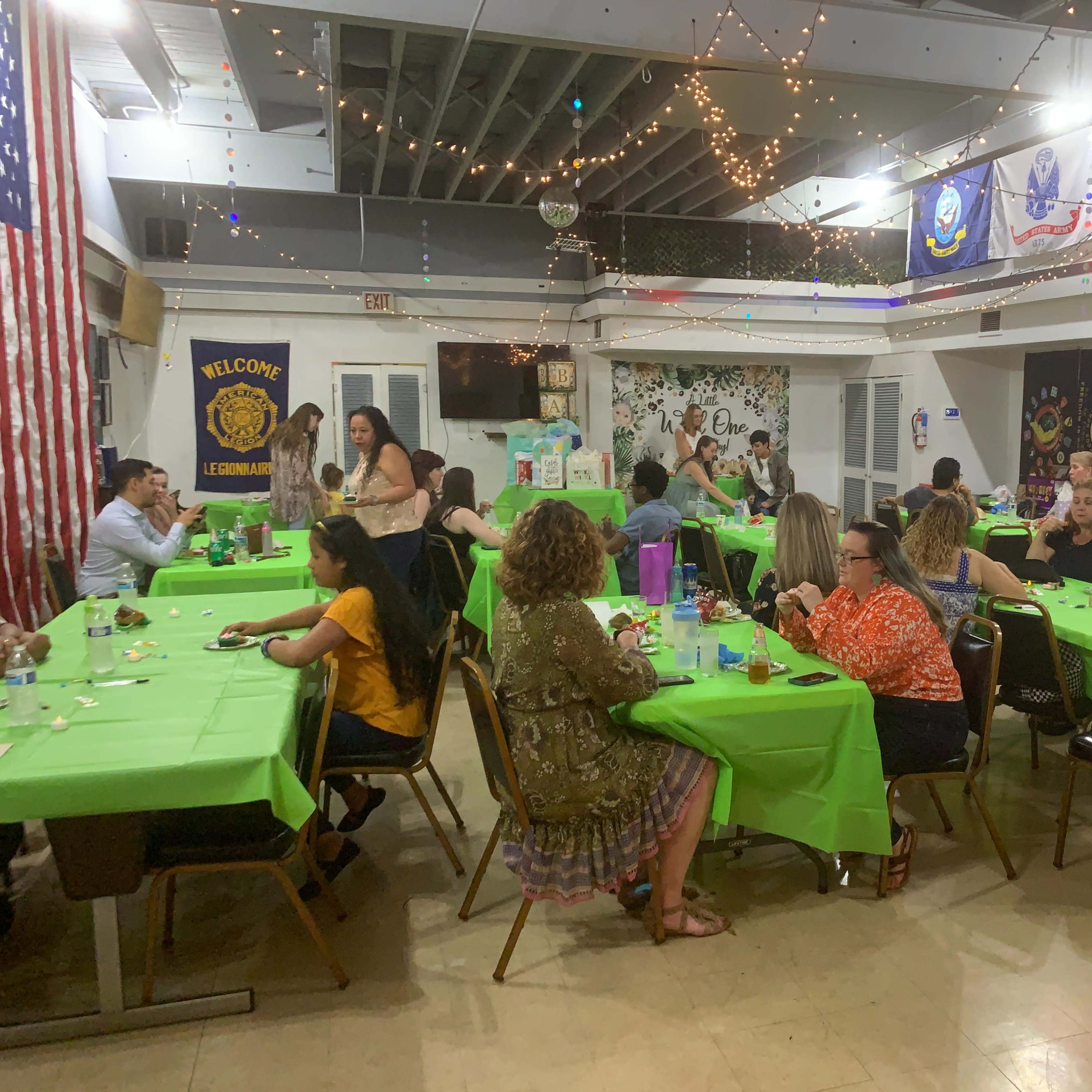 A group of people sits at tables covered with green tablecloths, enjoying a gathering in a decorated hall.