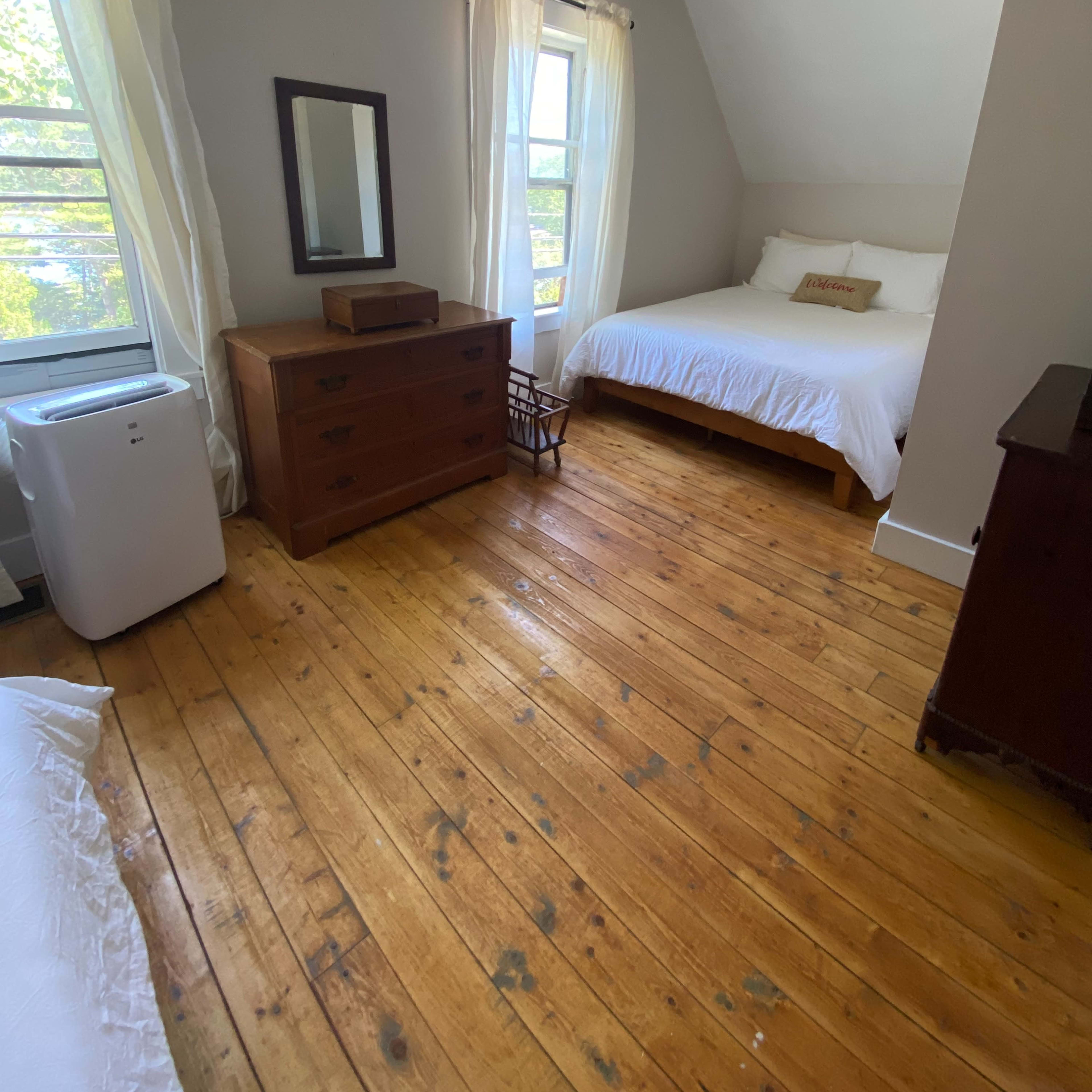 A bedroom with a wooden floor, a bed with white bedding, a wooden dresser, and two windows with sheer curtains.