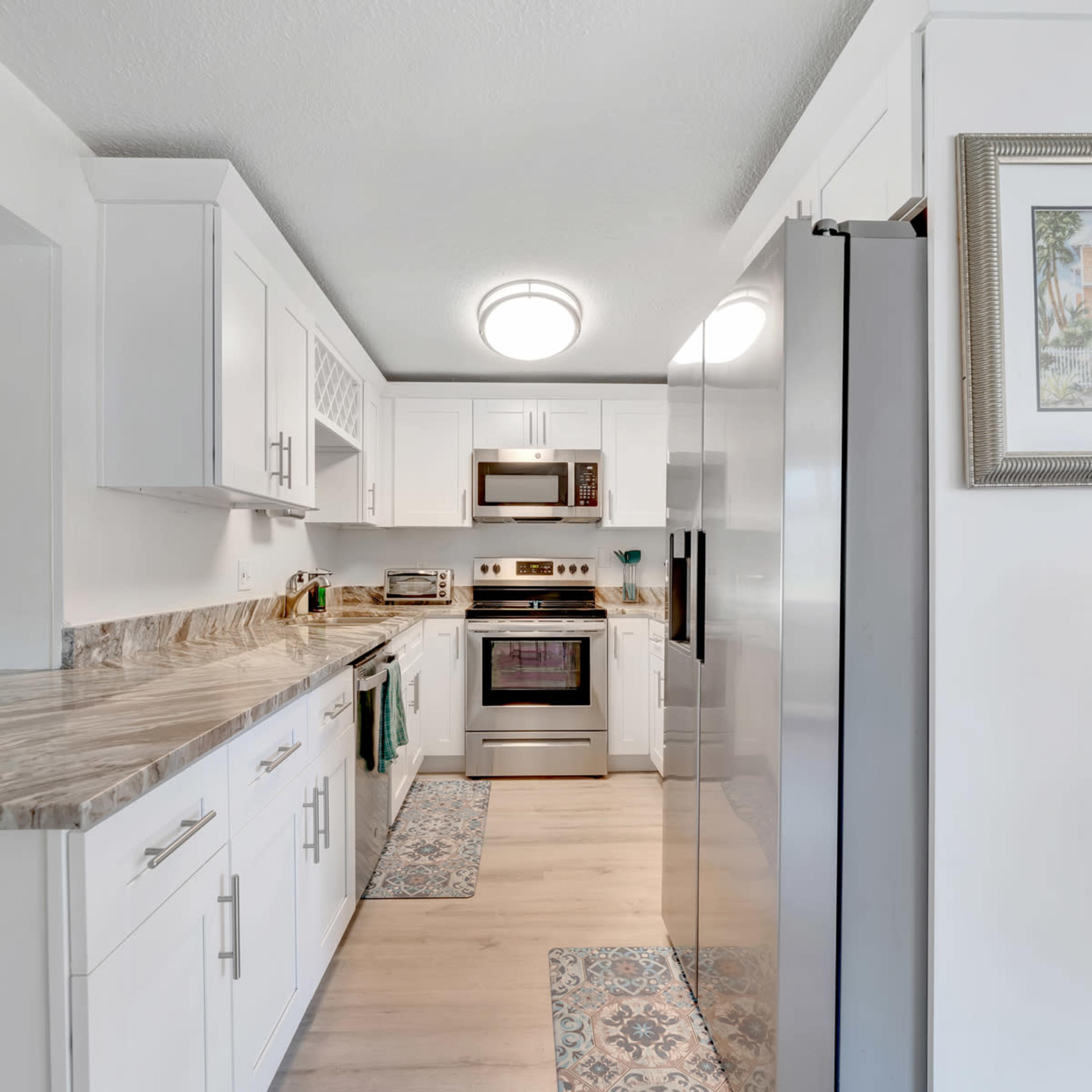 A well-lit kitchen features white cabinetry, stainless steel appliances, and a marble countertop with decorative rugs on a wooden floor.