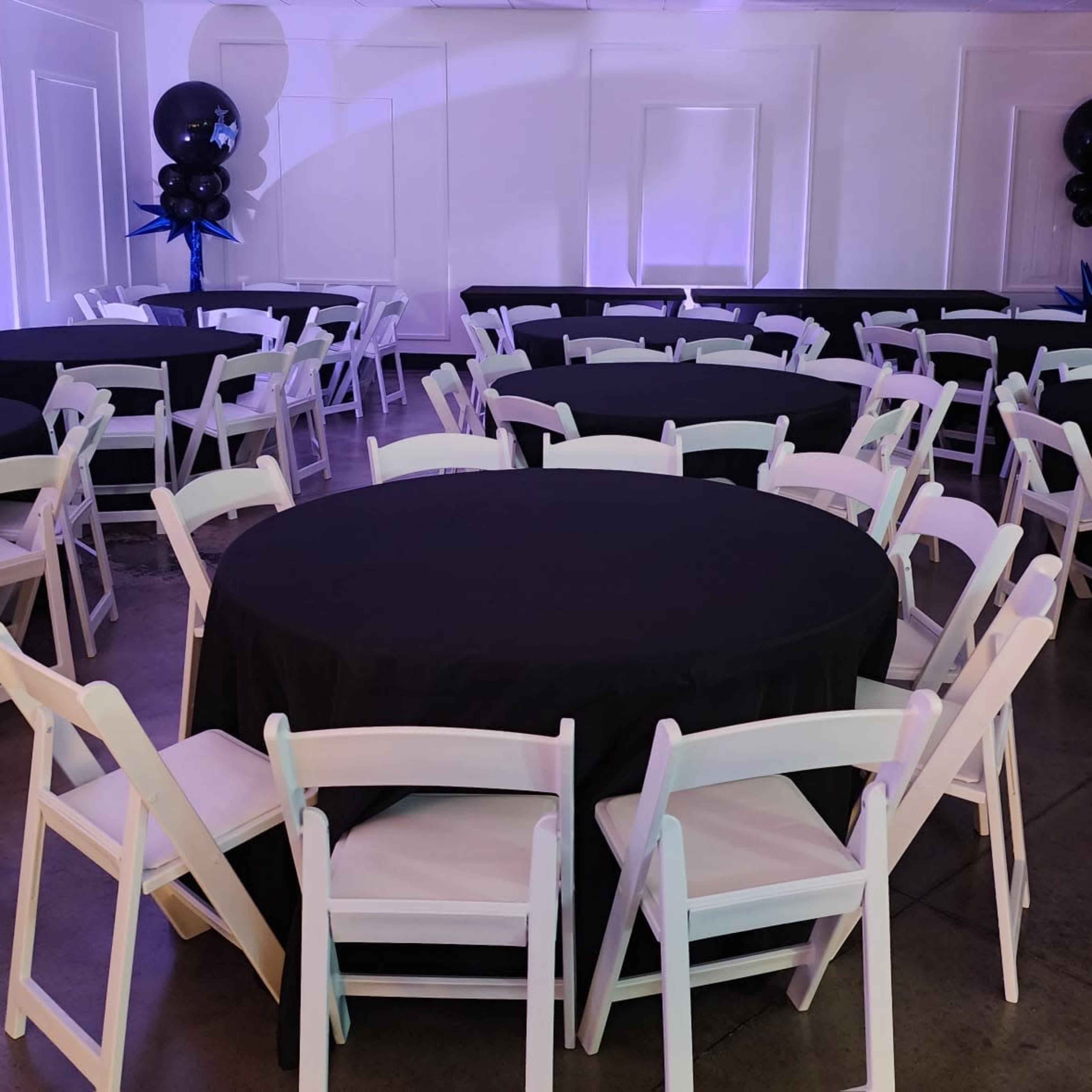 A room set up for an event, featuring several round tables covered with black tablecloths and surrounded by white folding chairs.