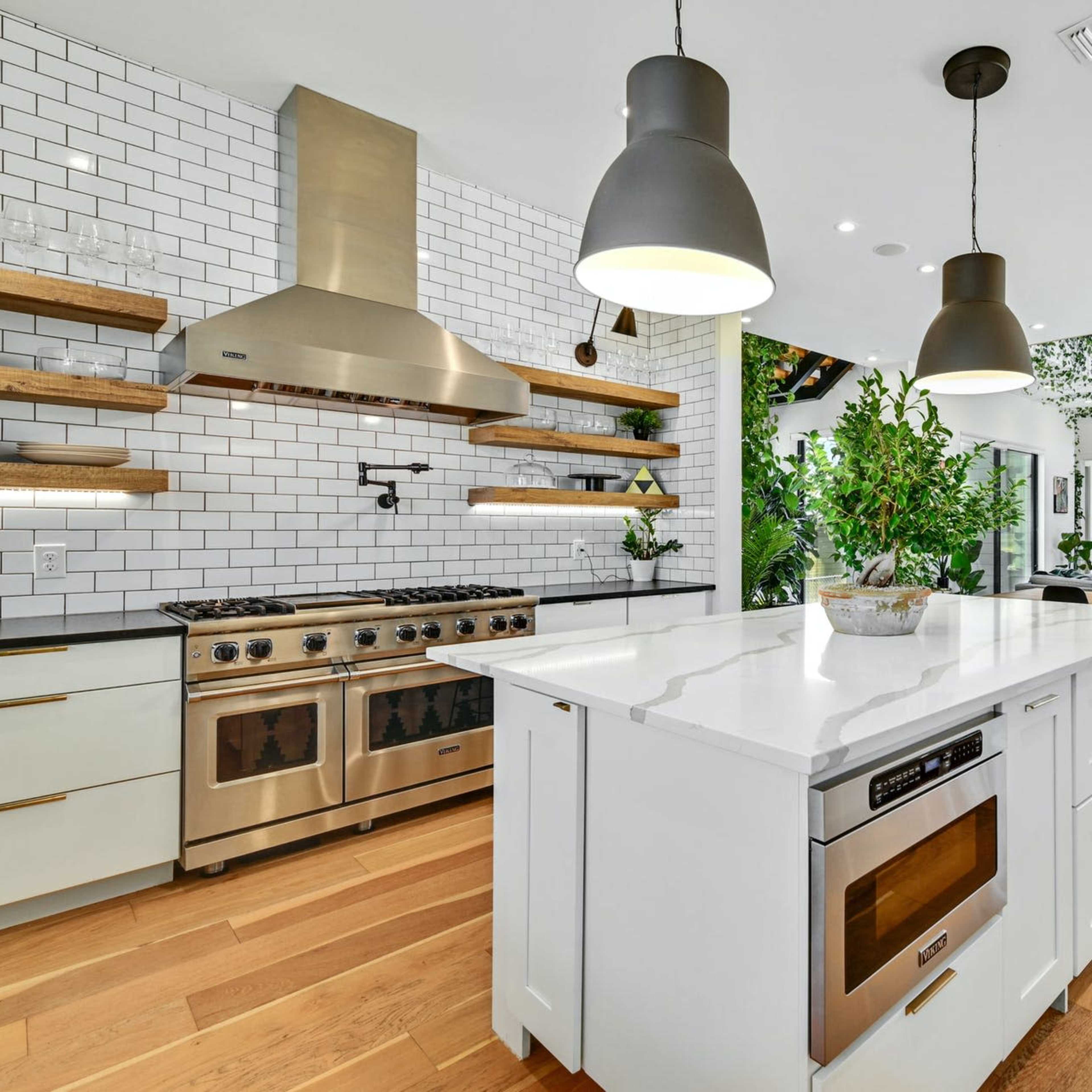 A modern kitchen features a central island with a stovetop and oven, surrounded by open shelving and large windows overlooking a living area adorned with plants.