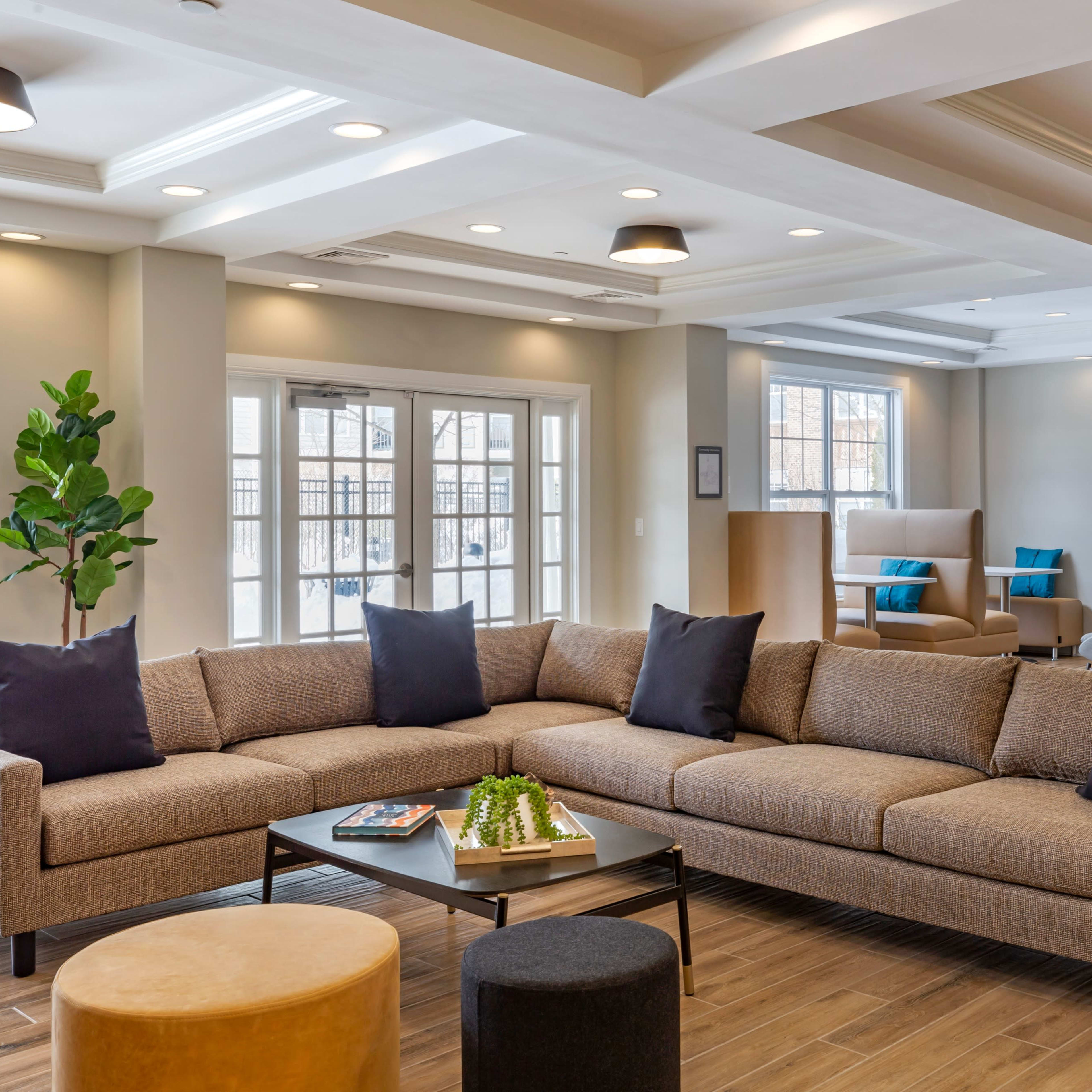 A modern seating area featuring a large sectional sofa with dark pillows, a coffee table, and a minimalistic bookshelf, all set in a well-lit room with large windows.