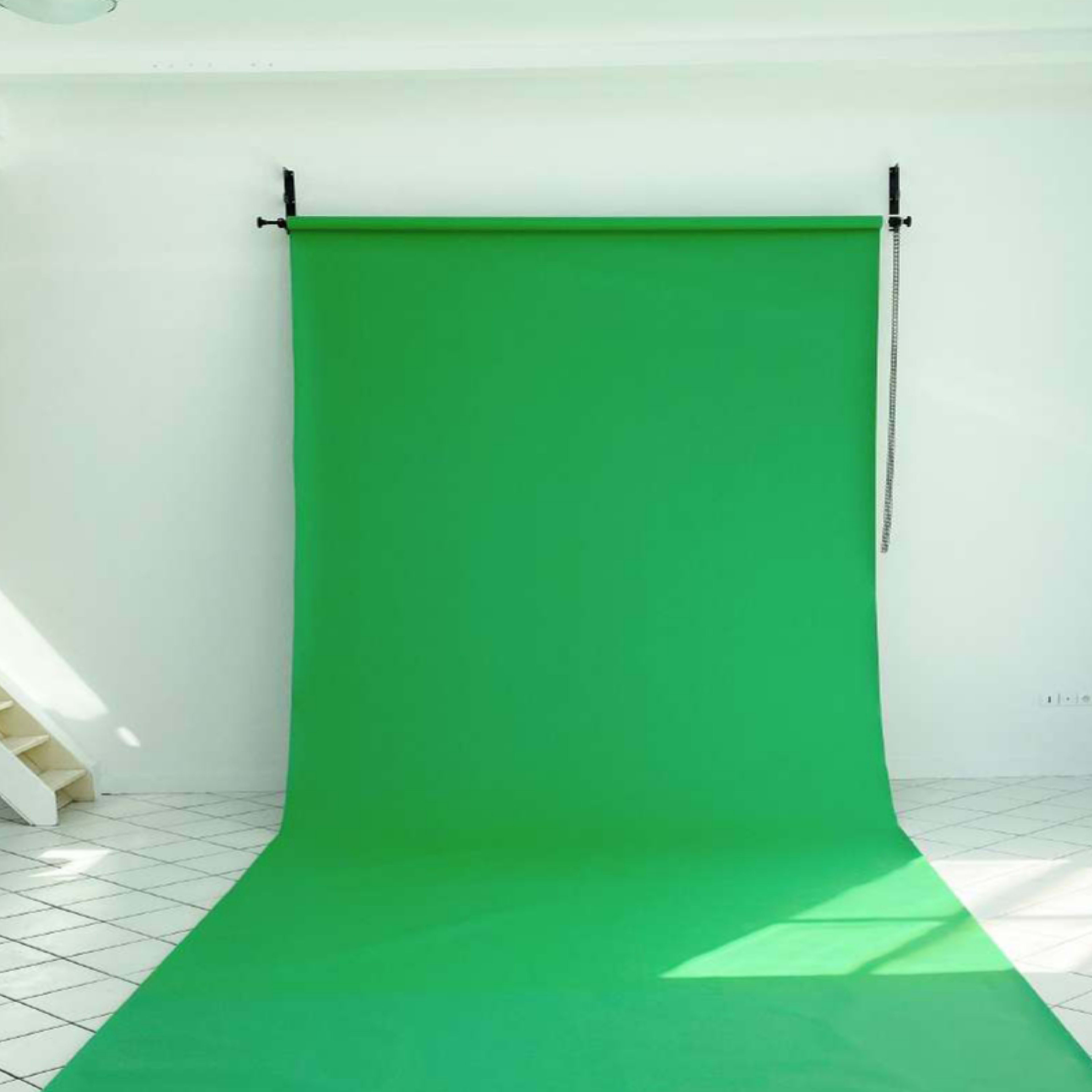 A green backdrop is set up in a brightly lit room with white walls and a staircase in the background.