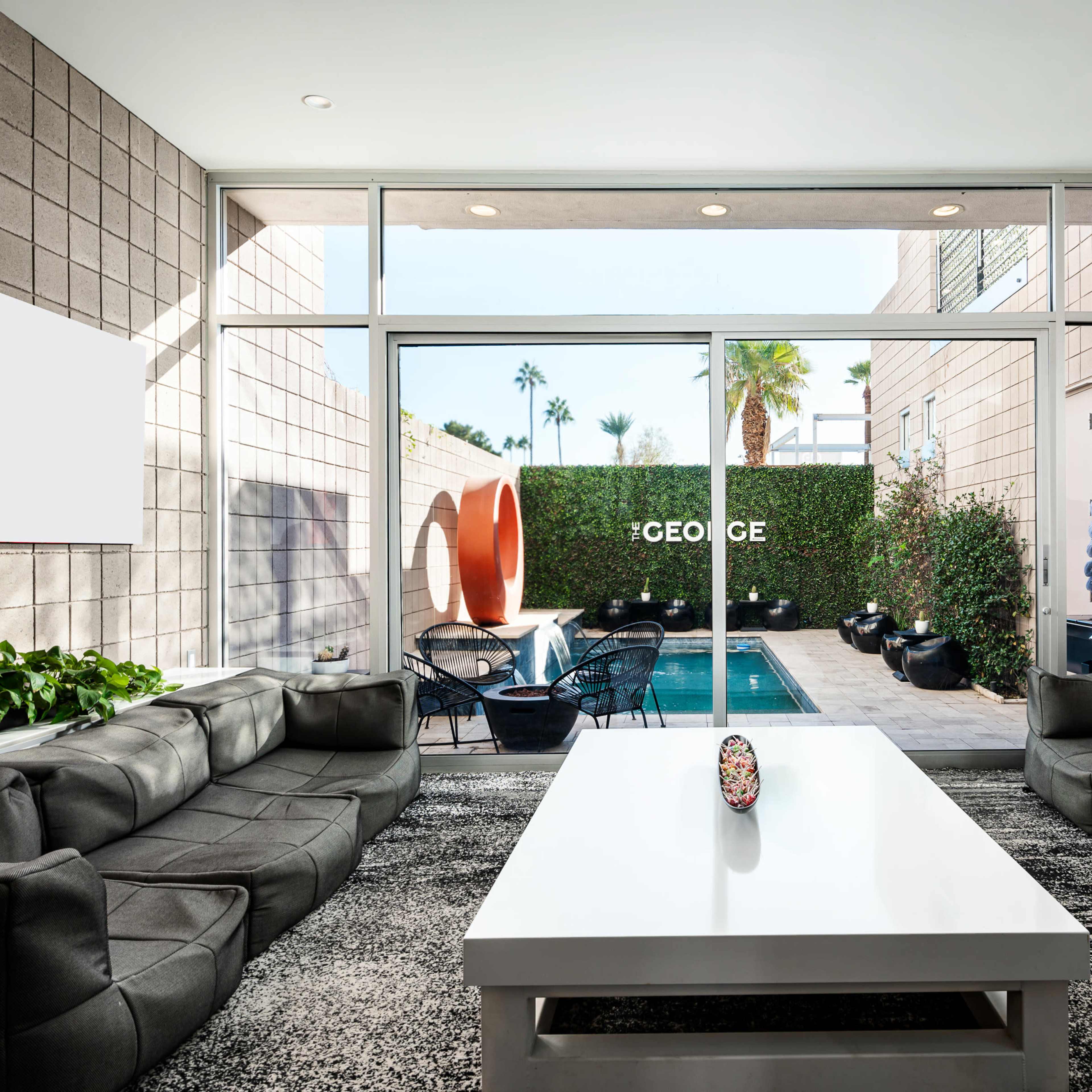 A modern living room with large windows overlooking a patio featuring outdoor seating and a pool surrounded by greenery.