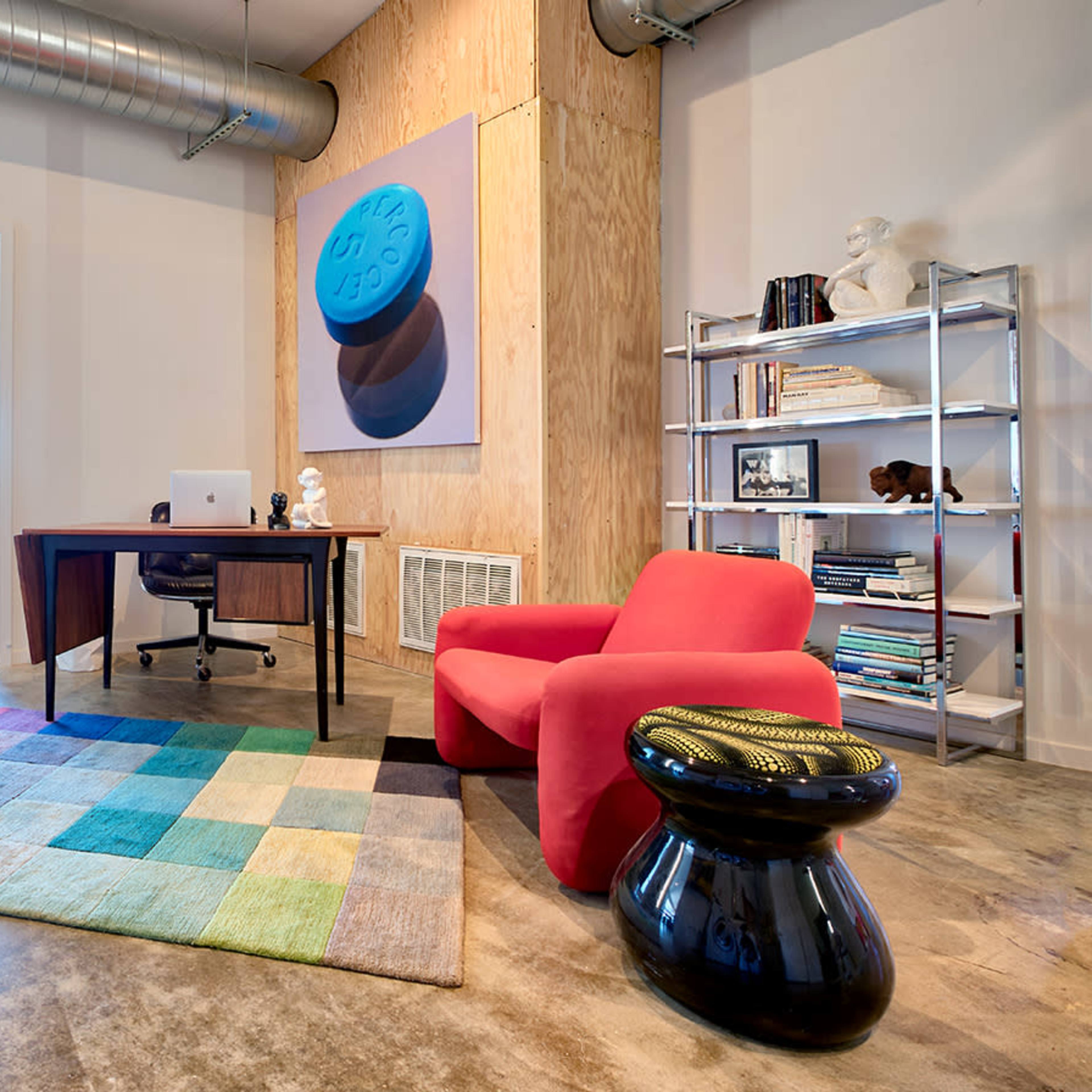 A modern office space featuring a wooden desk, a red chair, a patterned rug, and shelves filled with books and decorative items.