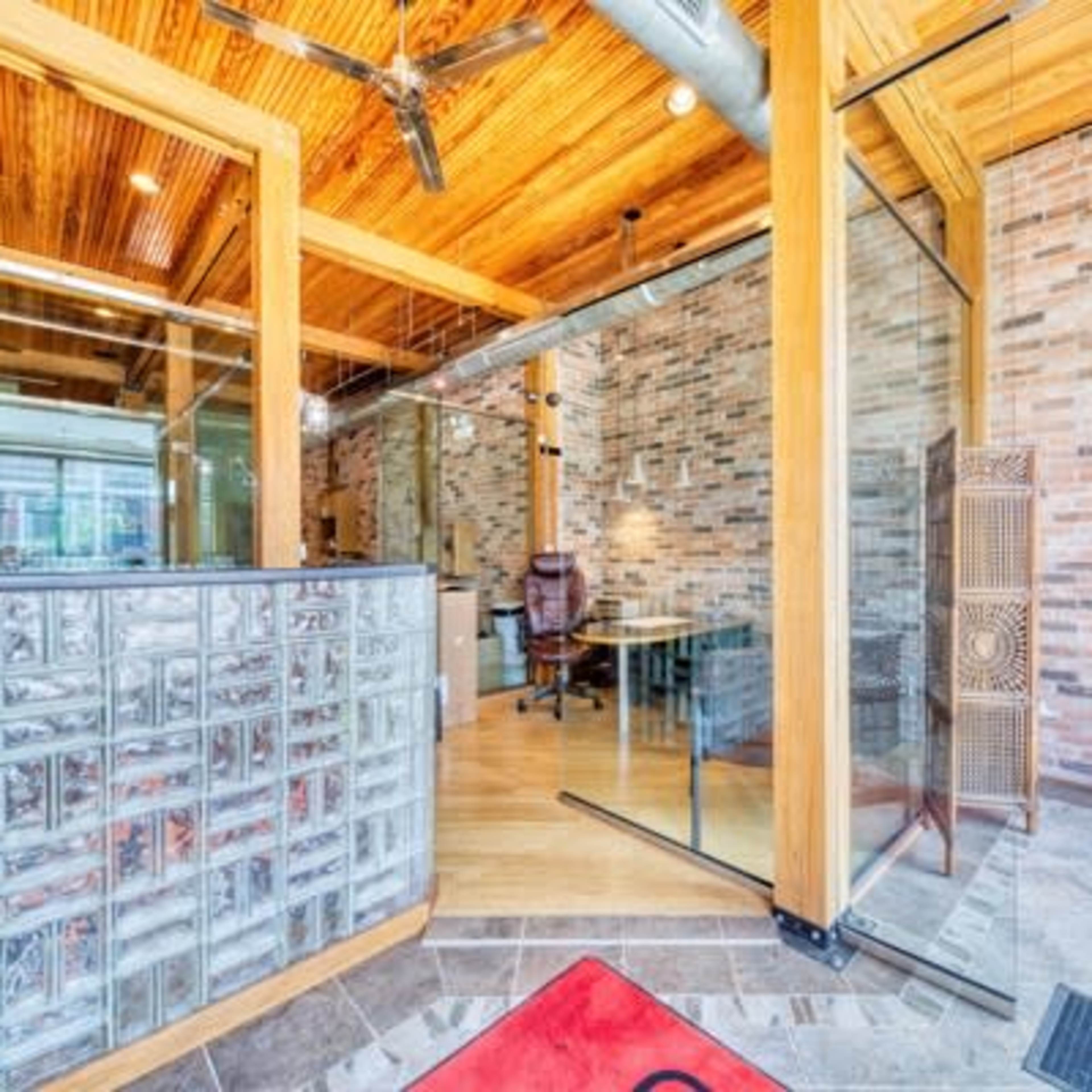 A modern office space features wooden ceilings, a glass-block wall, and a desk with a chair, set against a backdrop of exposed brick.