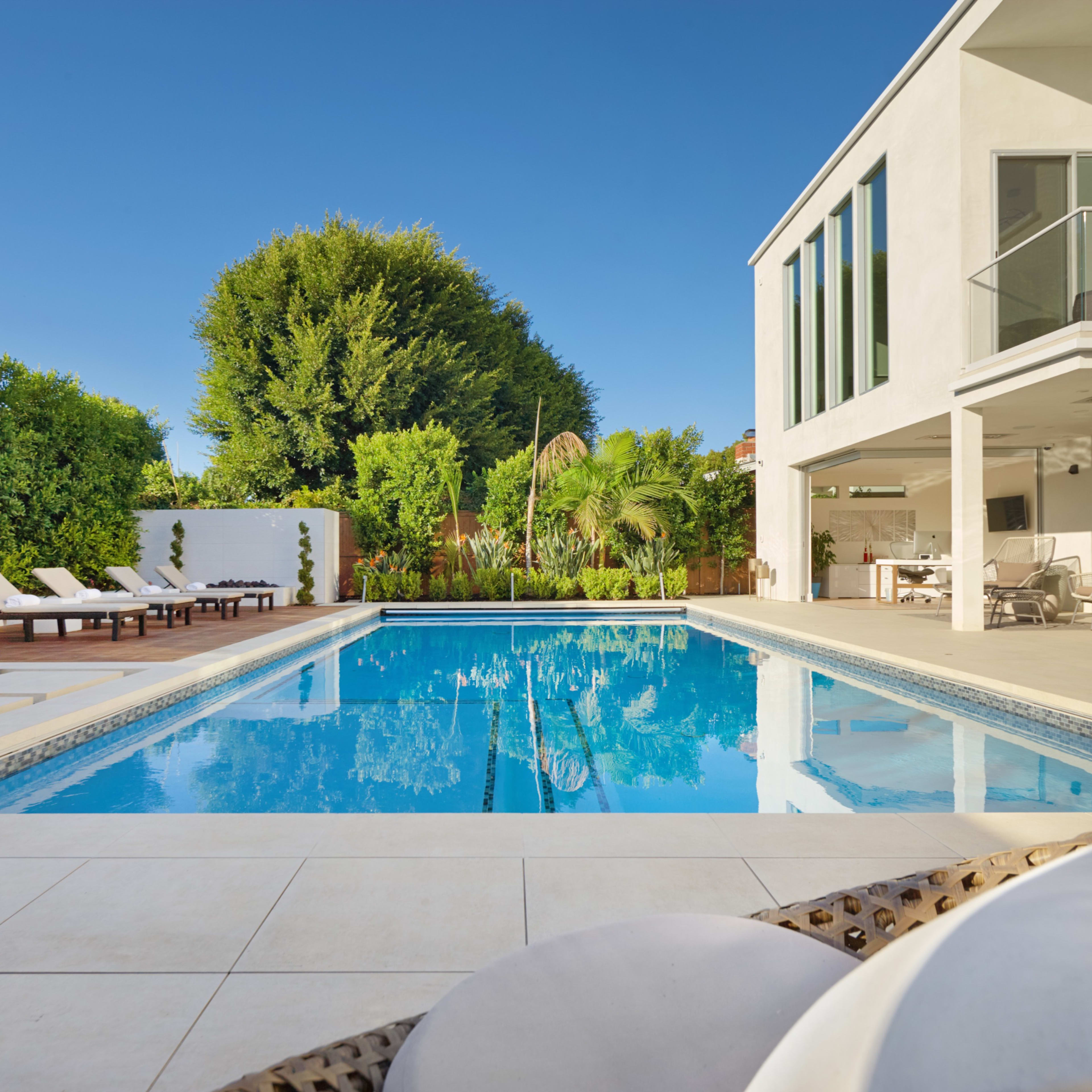 A modern backyard pool area features a large rectangular pool surrounded by lounge chairs and lush greenery.