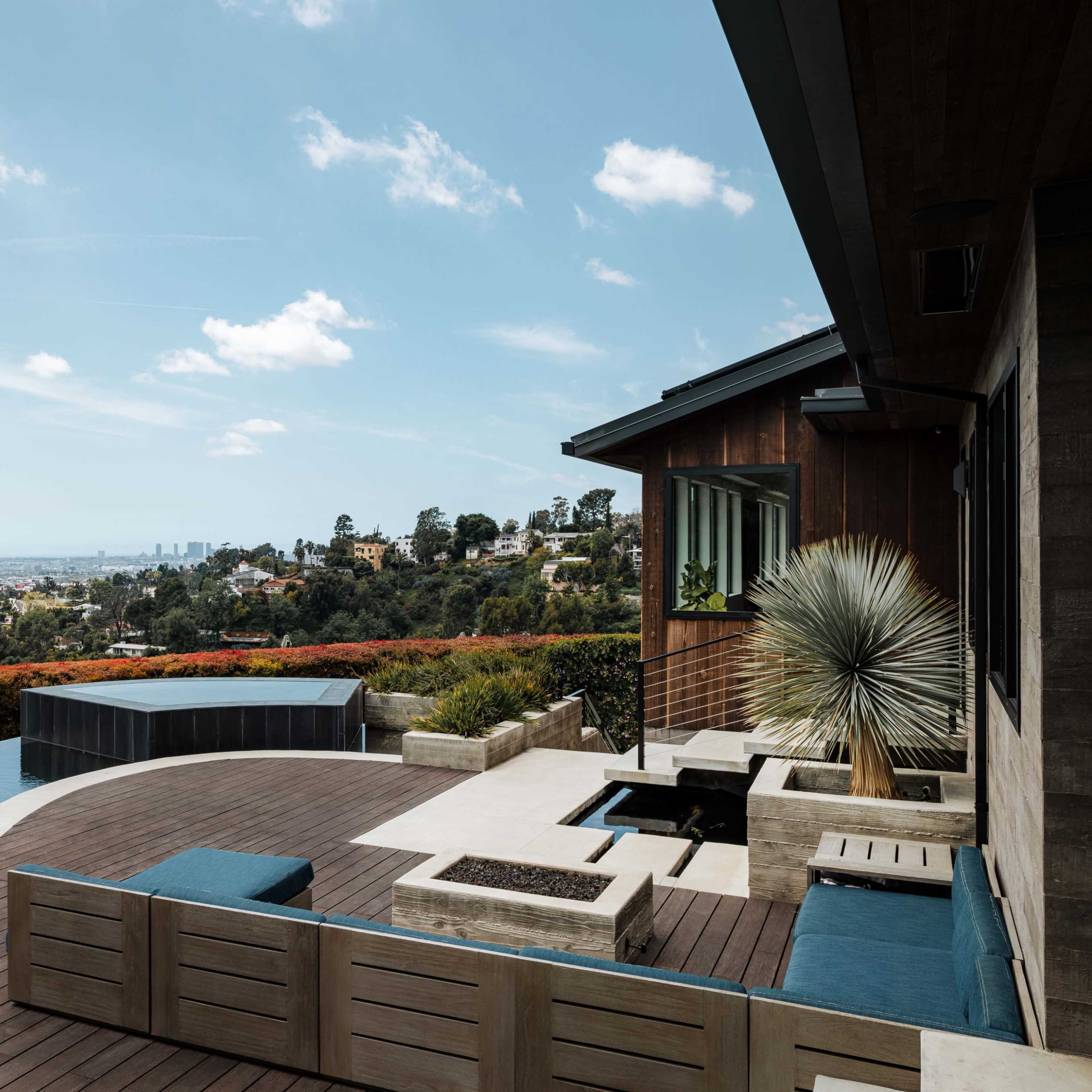 A modern outdoor living space with a wooden deck, a small pool, and stylish seating, overlooking a scenic view of a city and surrounding hills.