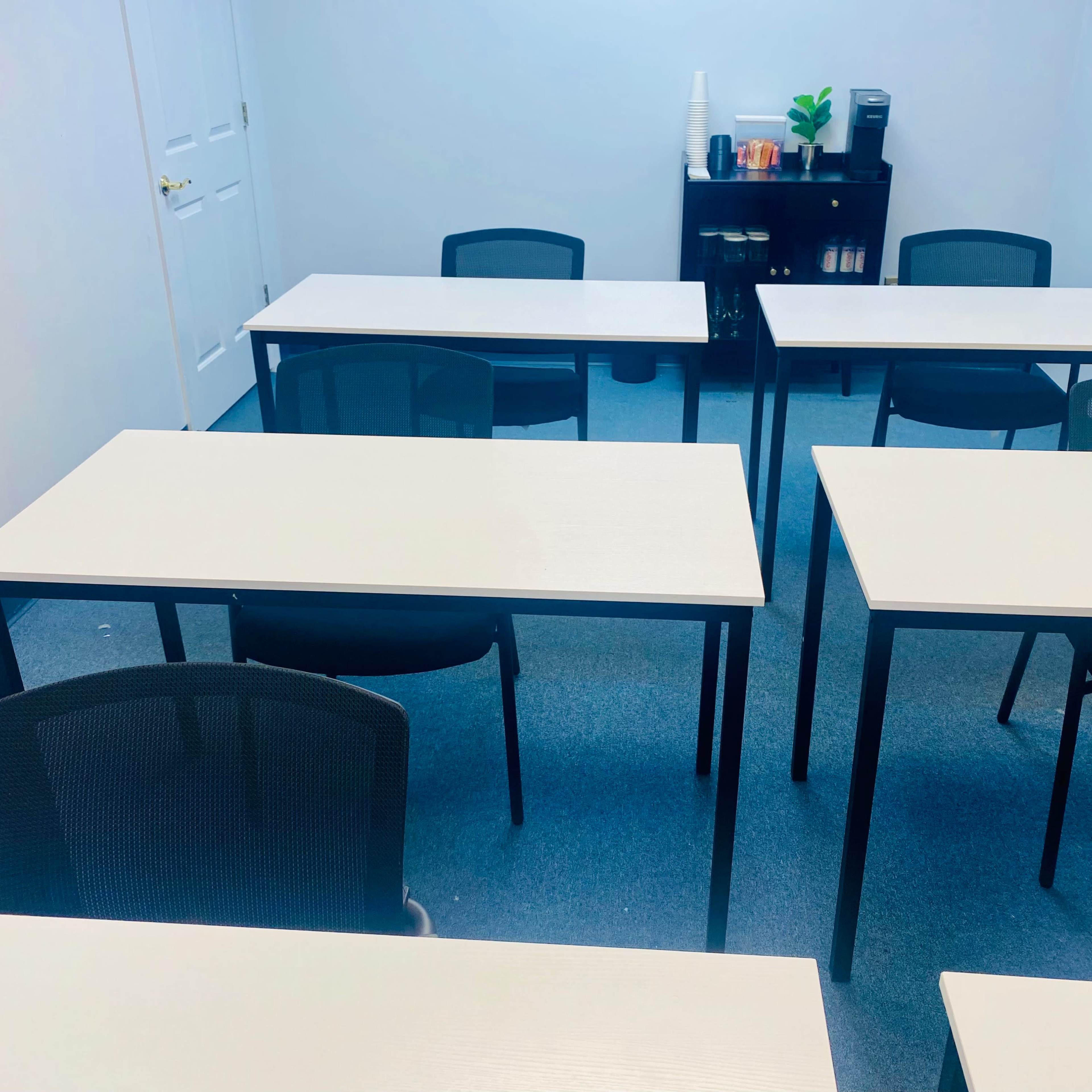 A brightly lit classroom with several empty tables and chairs arranged neatly on a blue carpeted floor, alongside a small shelf holding various items.