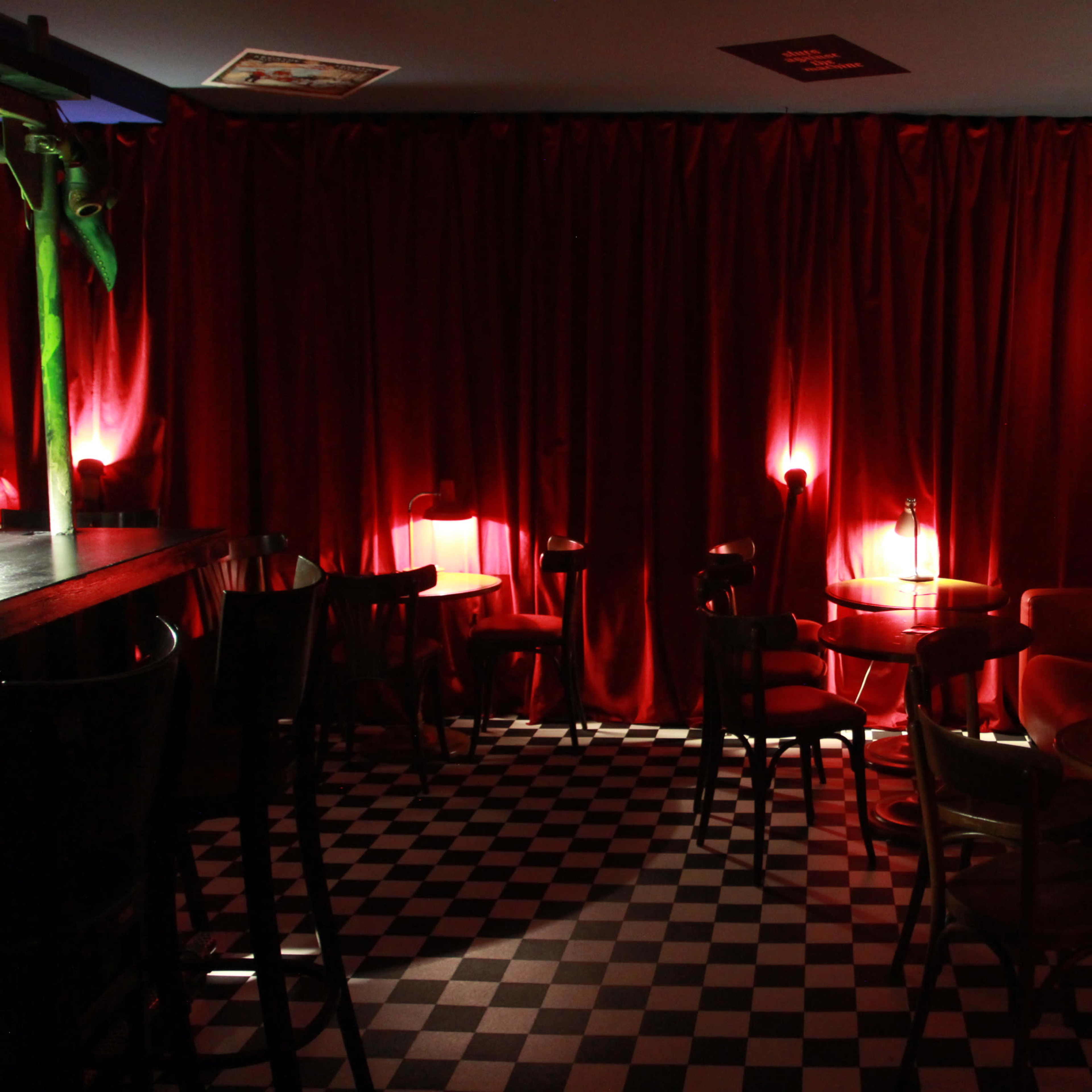 The interior of a dimly lit bar features red curtains, checkered flooring, and small tables illuminated by soft lighting.