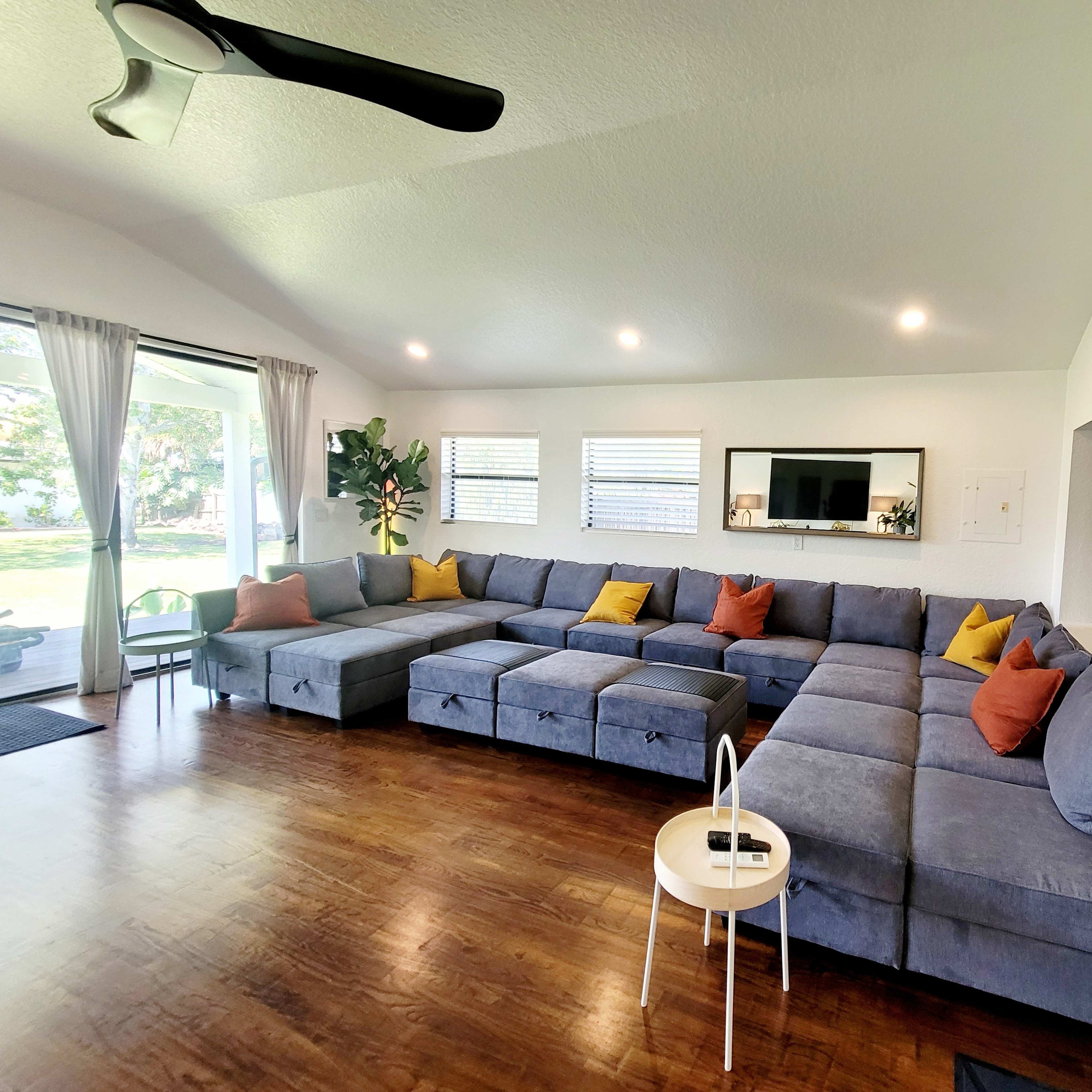 A spacious living room with a large sectional couch adorned with orange and yellow pillows, a small white table, and large windows letting in natural light.