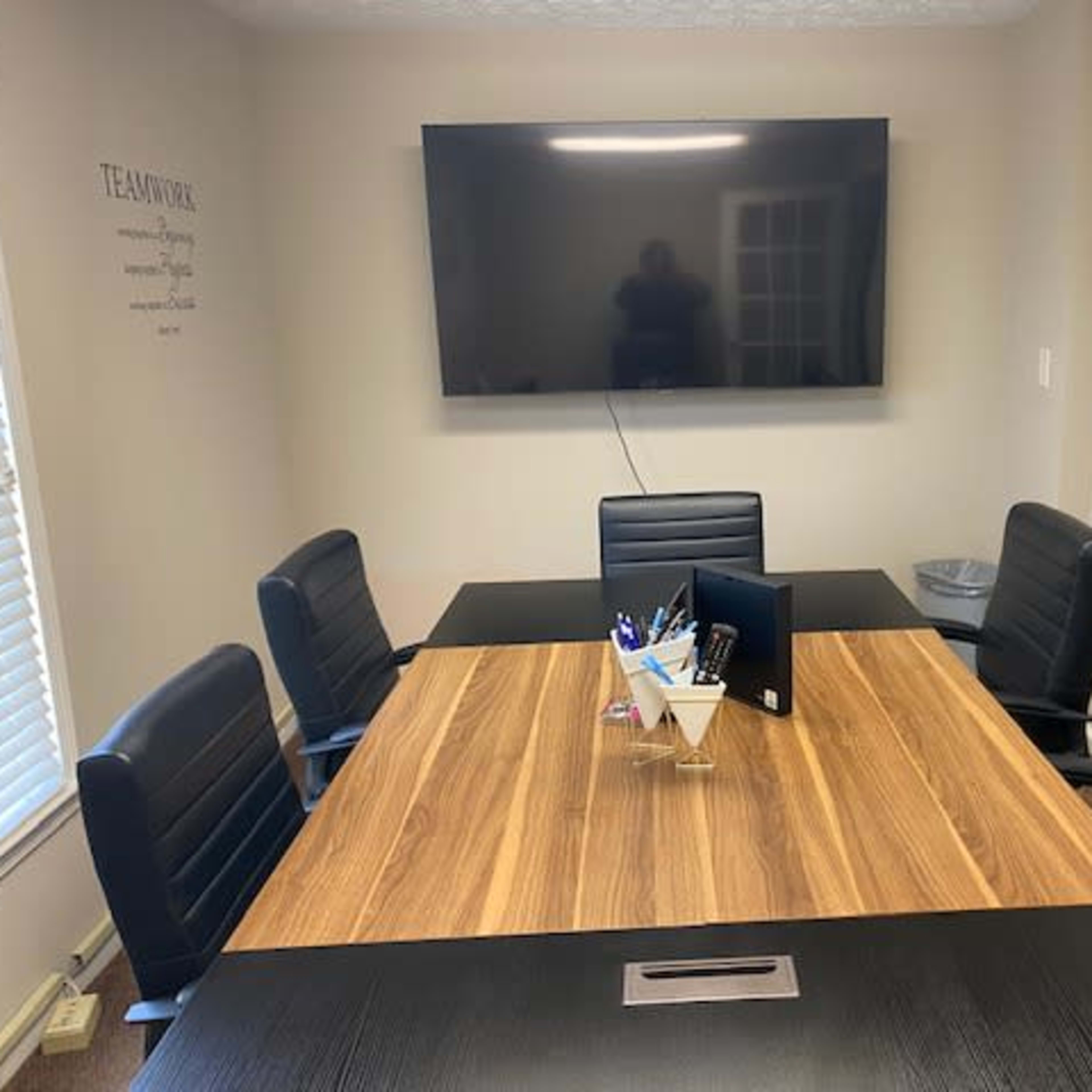 A conference room features a large wooden table surrounded by black chairs, a wall-mounted TV, and a whiteboard.